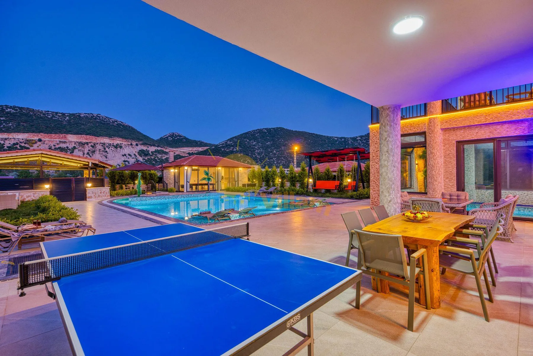 An outdoor poolside dining area with a blue table tennis table and a wooden dining table.