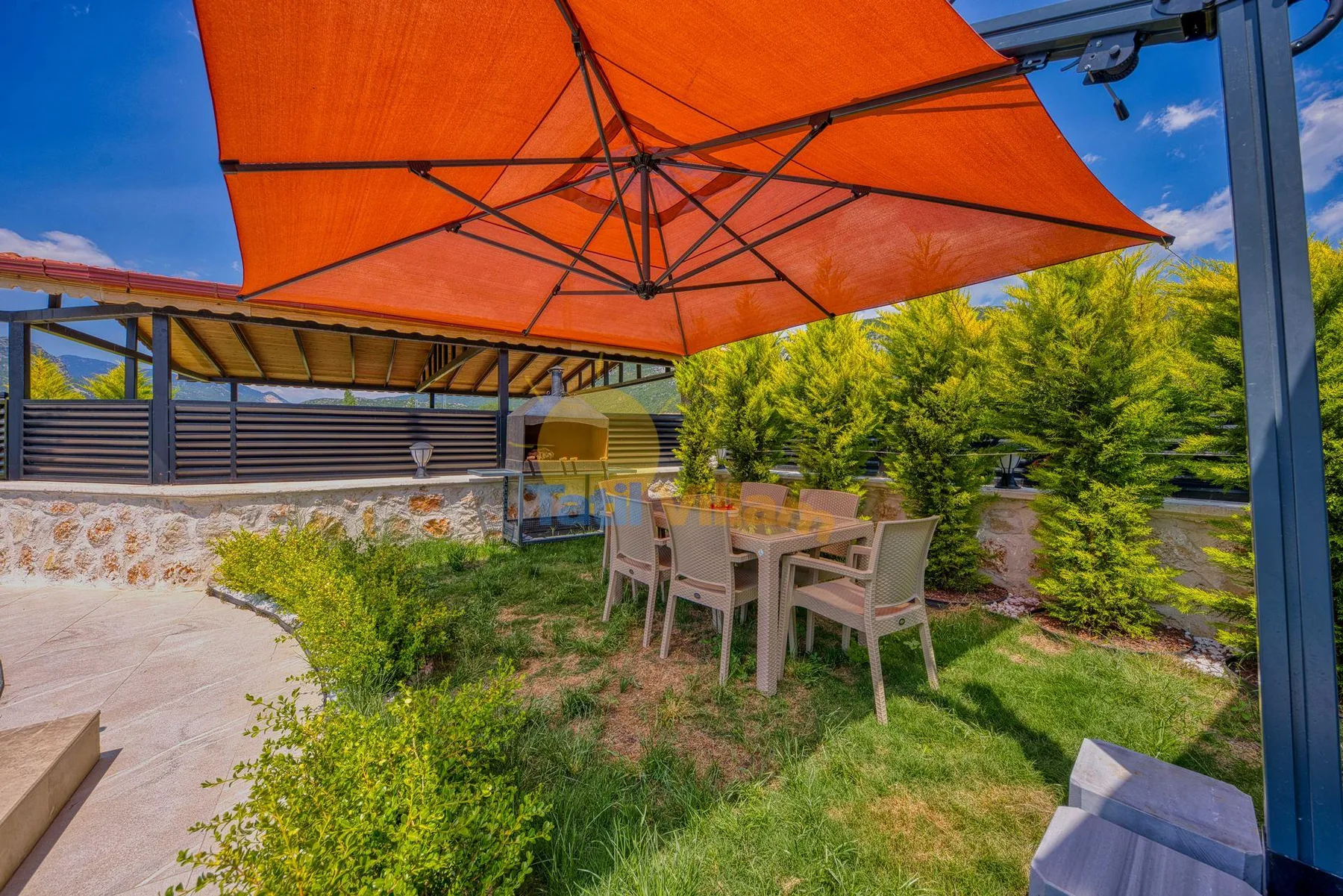 An outdoor dining area sits beneath a large orange shade canopy beside green shrubs.