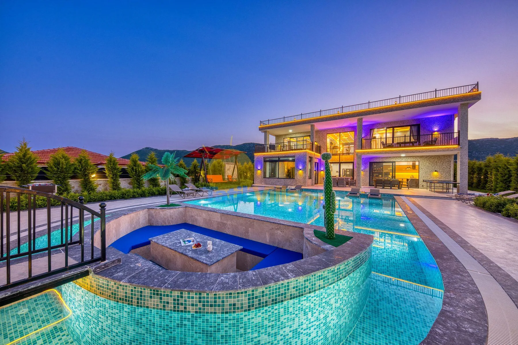 An illuminated modern villa overlooks a swimming pool and raised spa area.