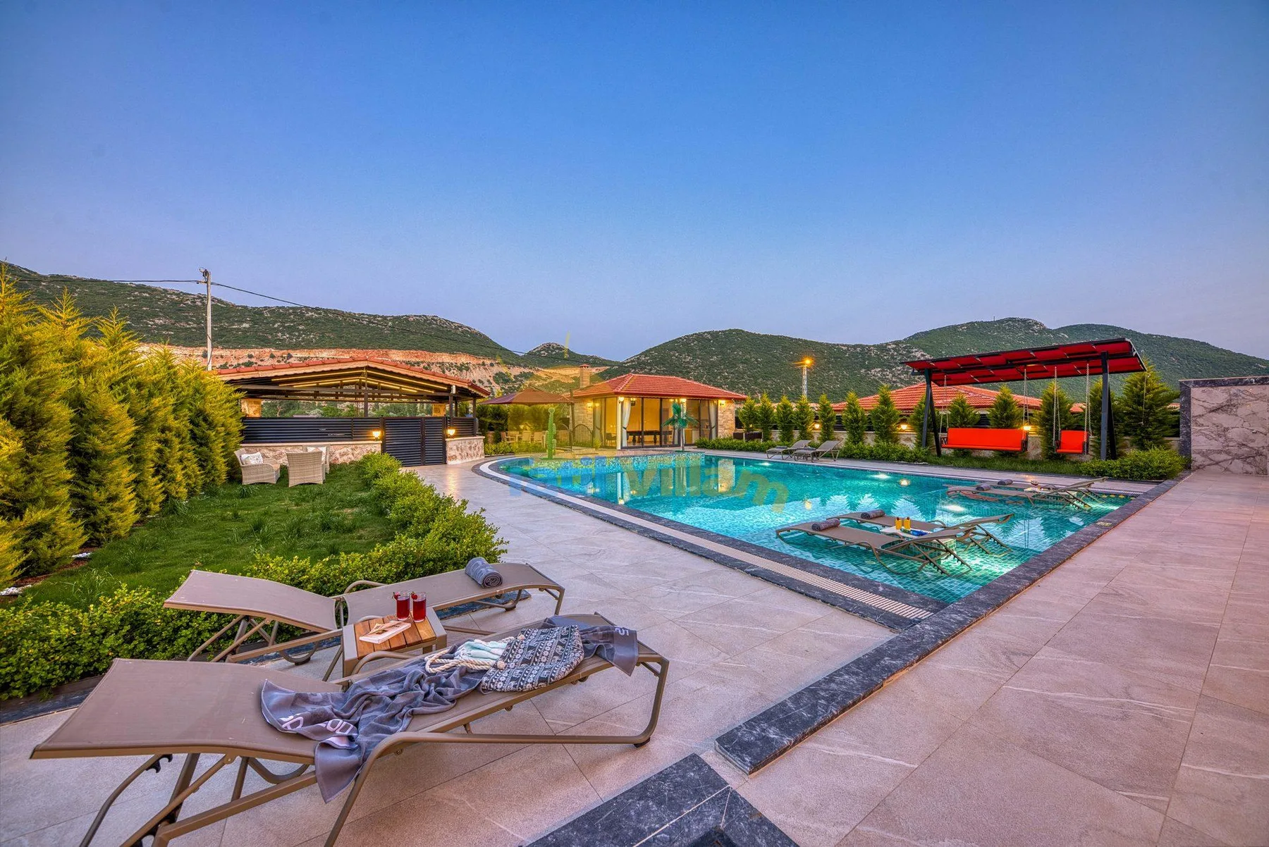 An outdoor swimming pool sits beside a paved patio with lounge seating and green hills beyond.