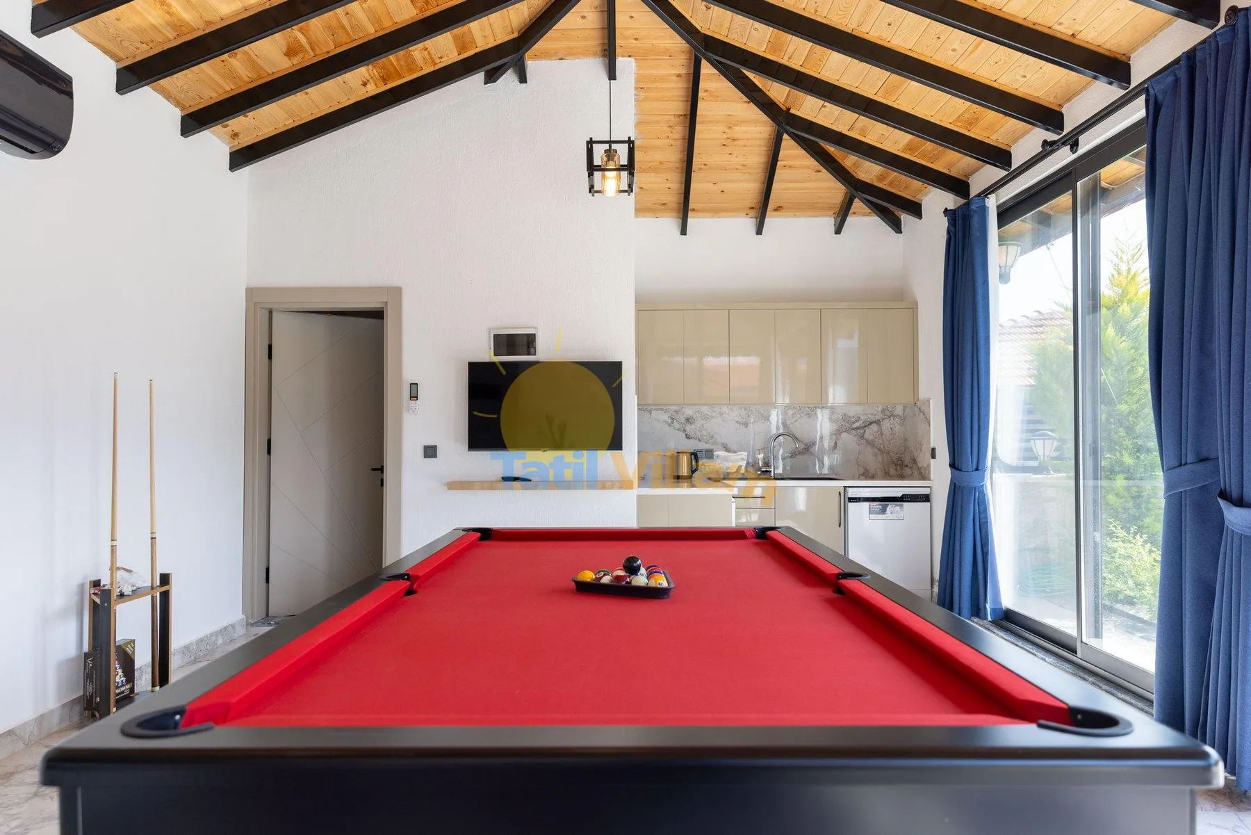 A billiards table sits in a bright room with a kitchenette and large windows.
