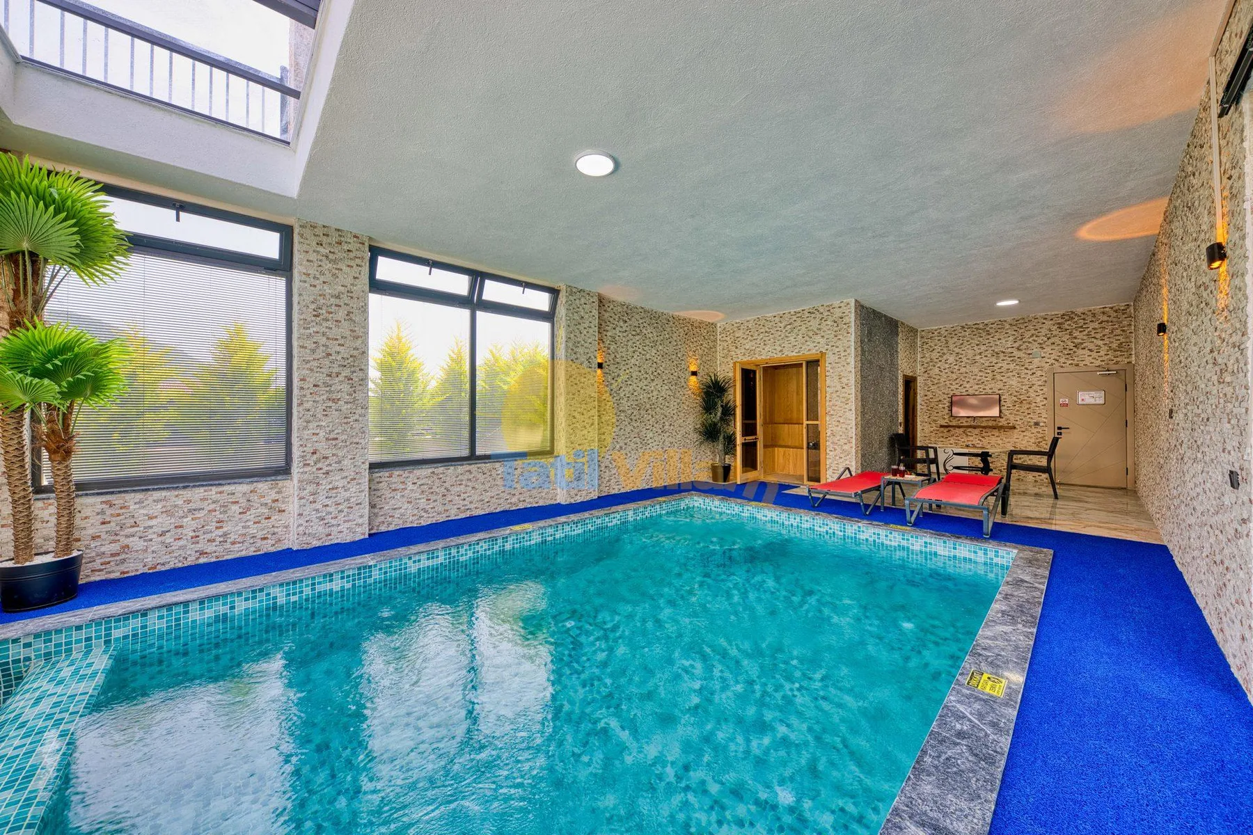 An indoor swimming pool fills a bright room with large windows and a blue deck.