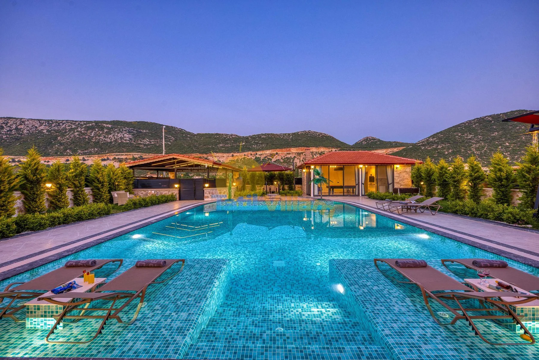An outdoor swimming pool with lounge chairs, landscaped grounds, and hills in the background.