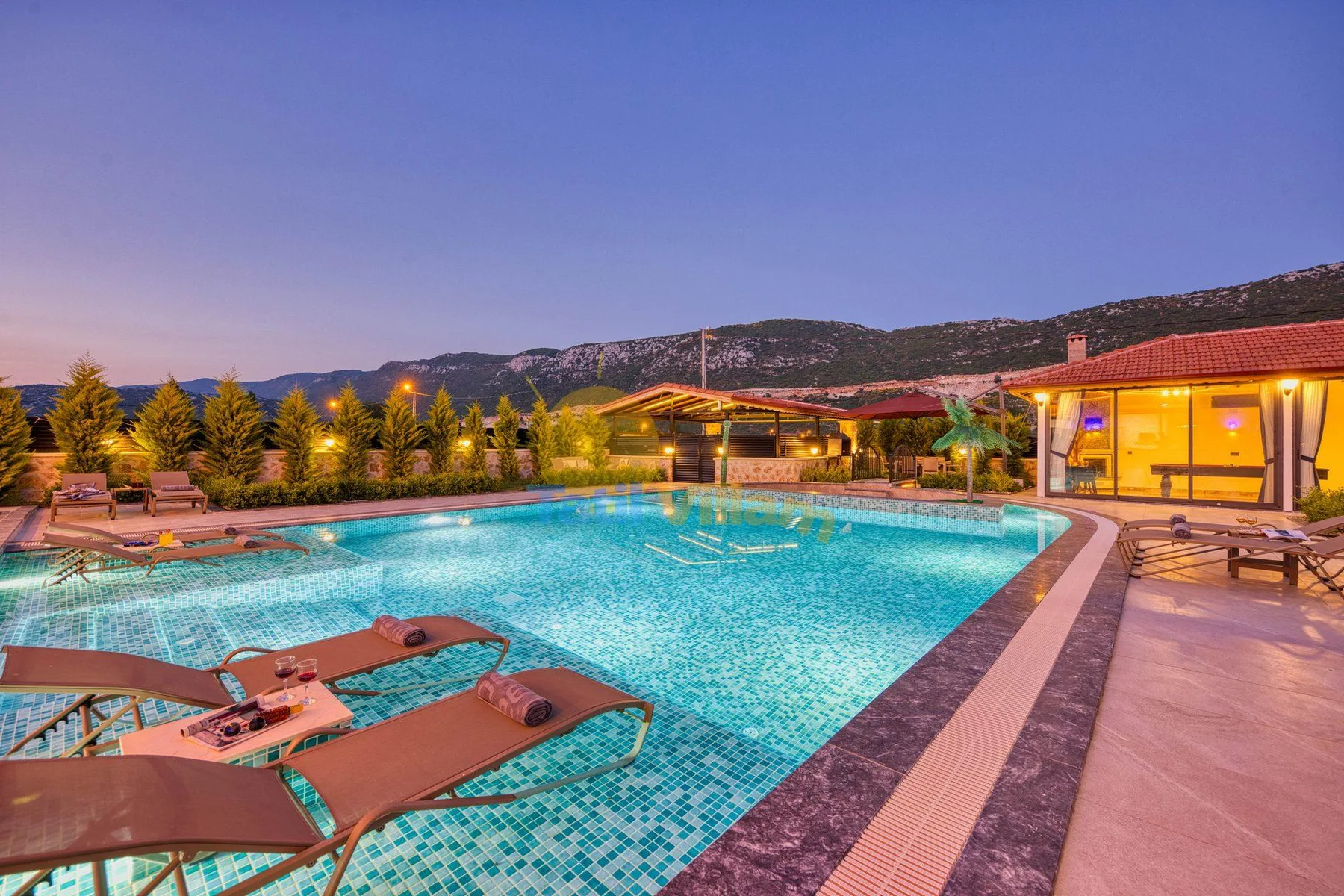 An outdoor swimming pool beside a lit building, with hills in the background.