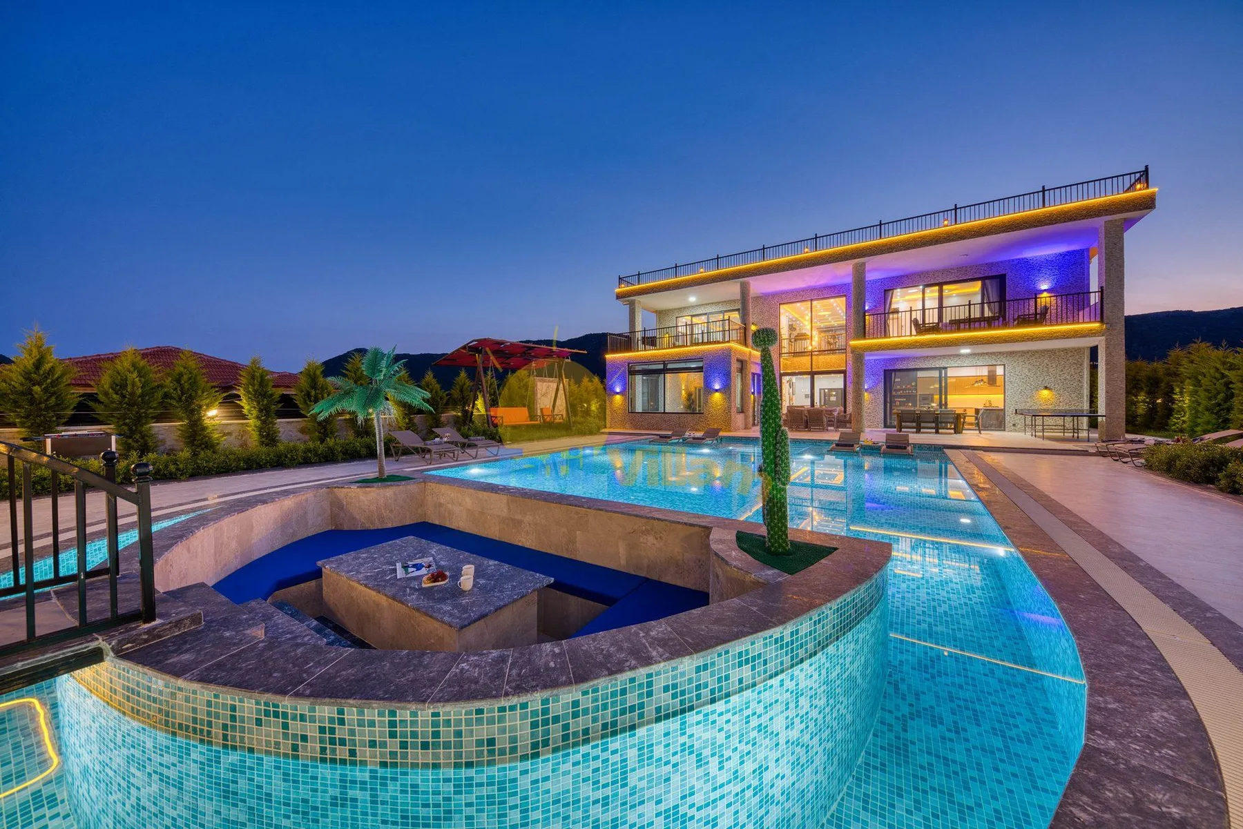 A modern house overlooks a brightly lit swimming pool at dusk.
