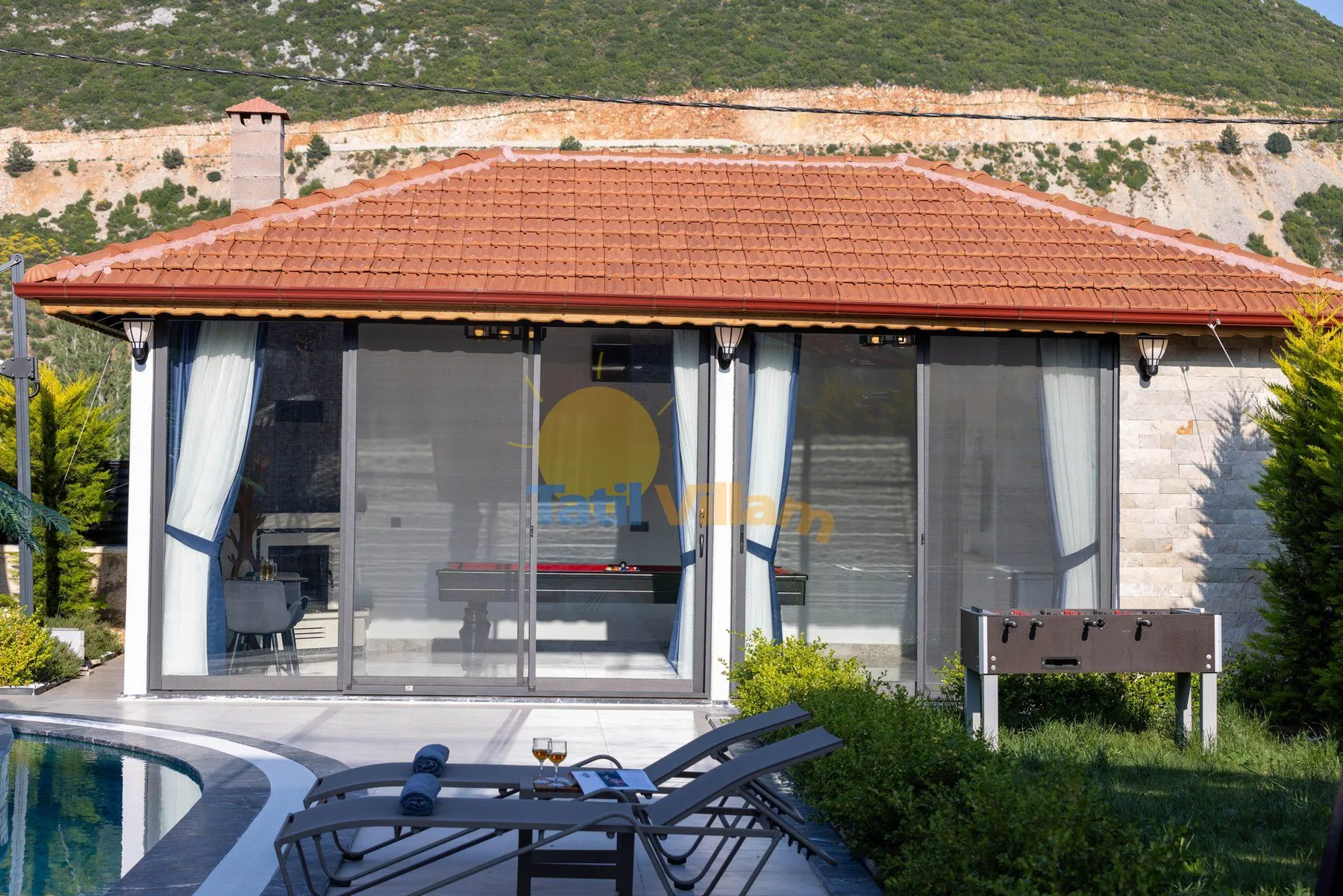 A glass-fronted poolside room with outdoor loungers and a table.