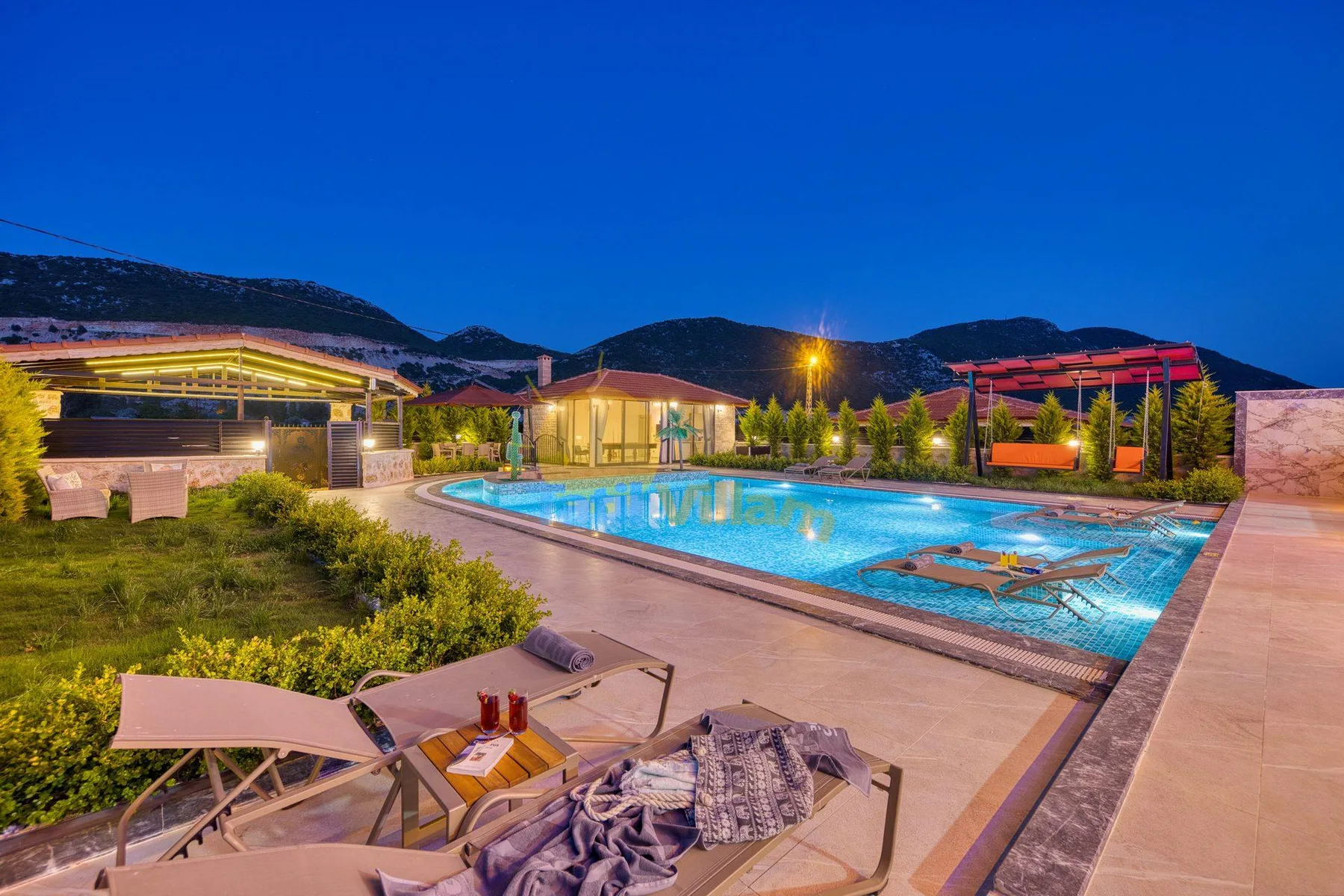 An outdoor swimming pool is surrounded by paved seating areas and landscaped grounds at dusk.