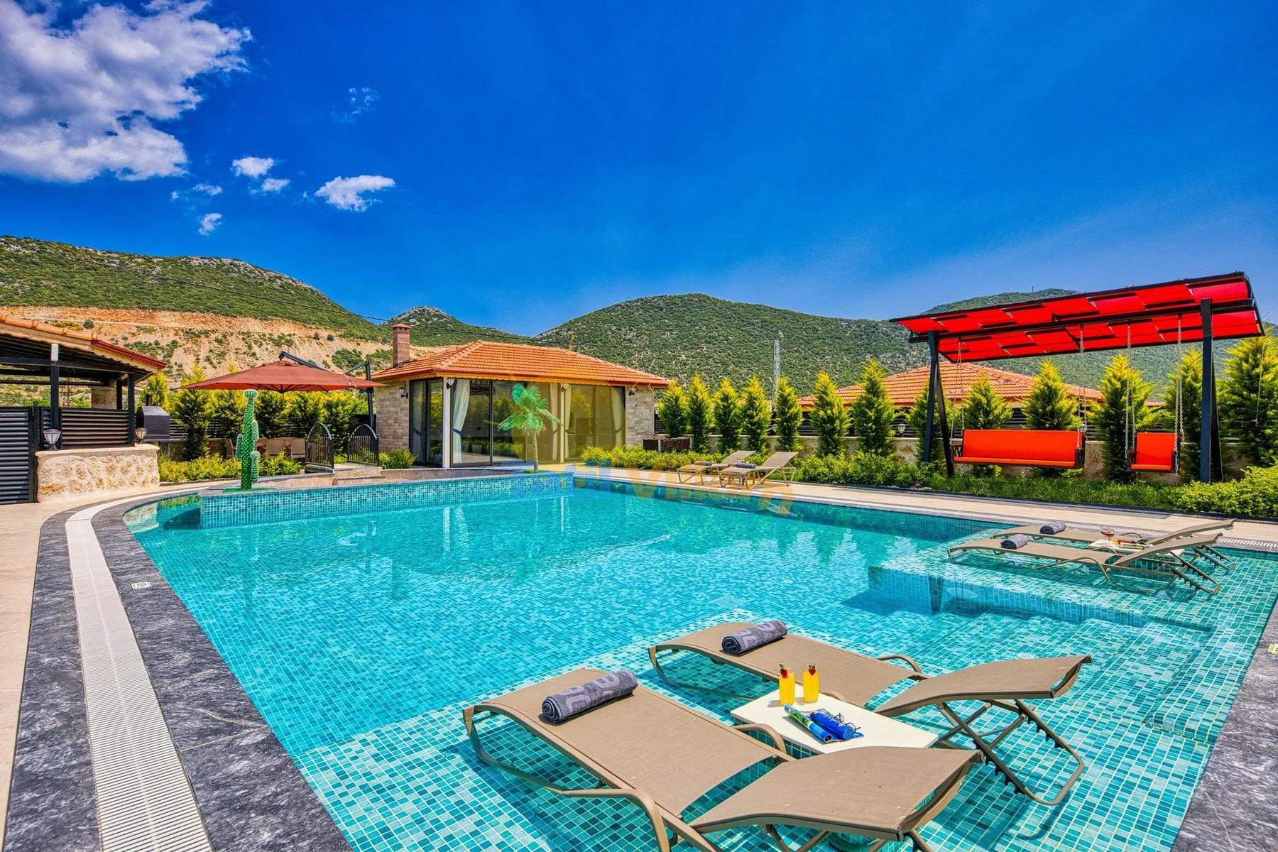 An outdoor swimming pool sits beside a low building, with hills in the background.