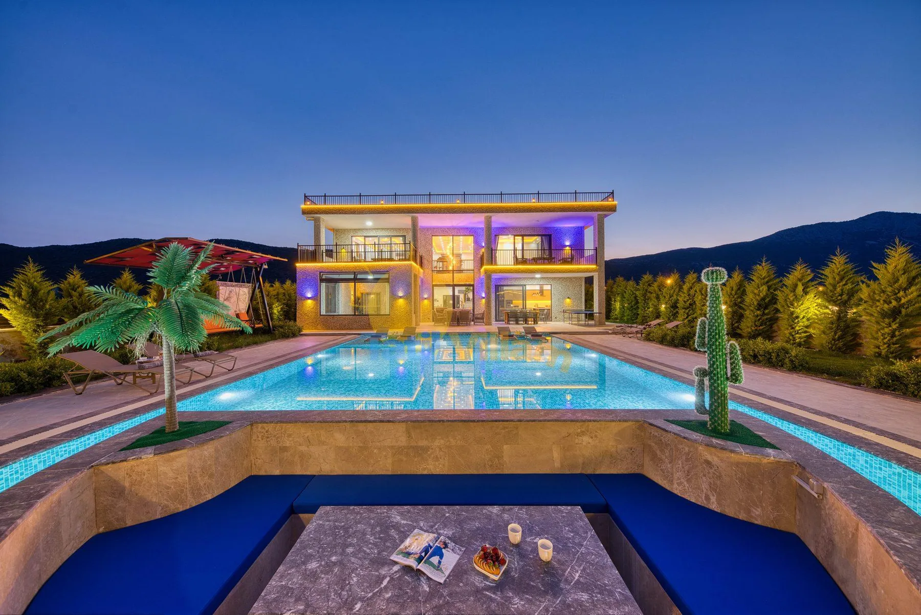 A modern house and illuminated swimming pool are seen at dusk, with mountains in the background.