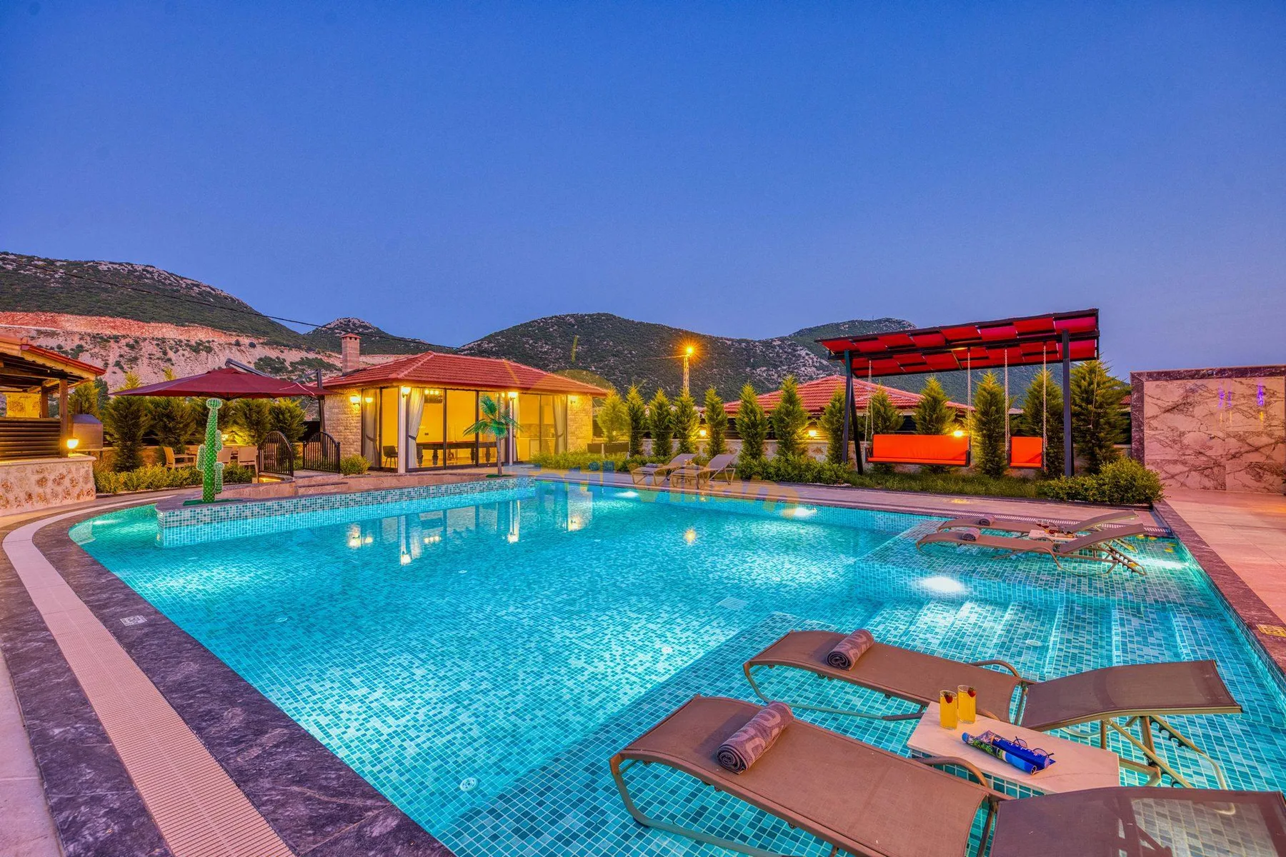A swimming pool with lounge chairs sits beside a warmly lit building.
