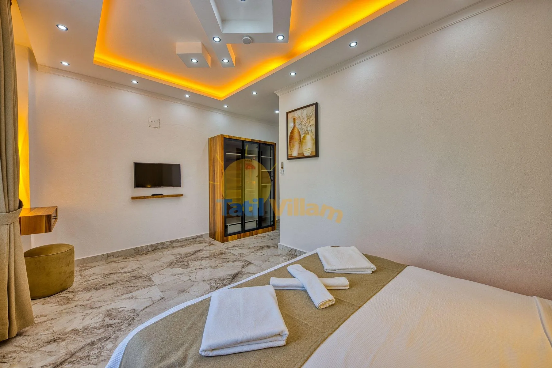 A bedroom with a bed, folded towels, a television, and warm recessed ceiling lights.