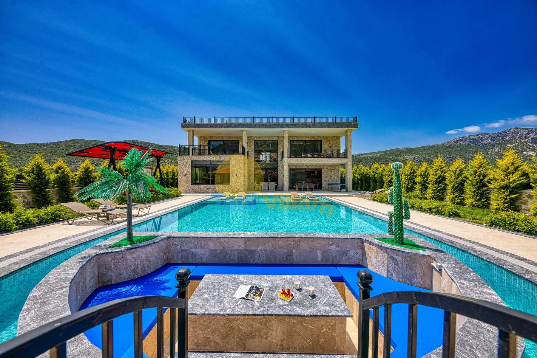 A modern house stands beyond a swimming pool, with mountains in the background.