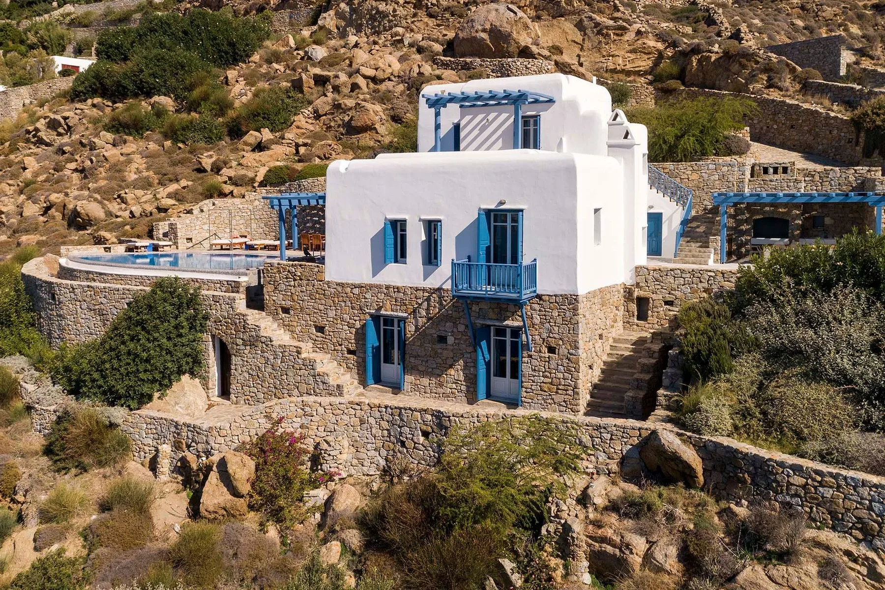 A white building with blue trim sits among stone walls on a rocky hillside.