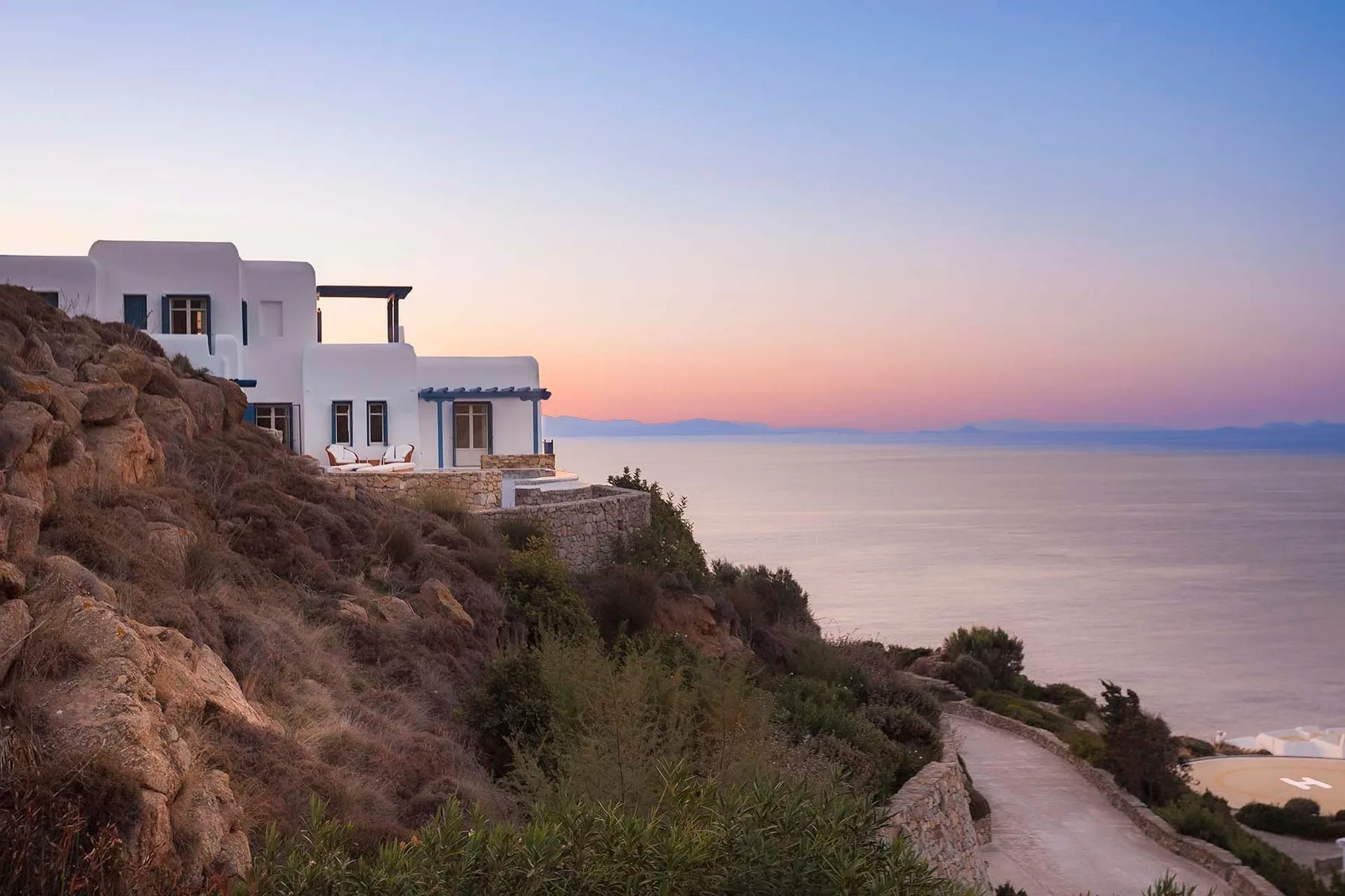 White buildings sit on a rocky hillside above the sea at sunset.