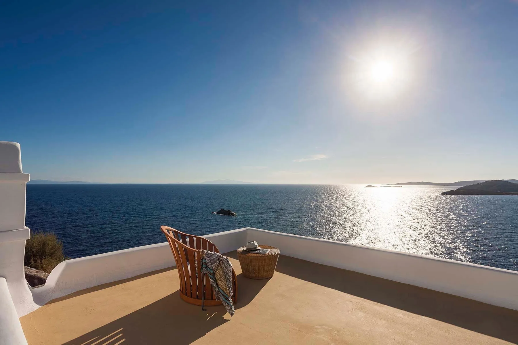 A seaside terrace overlooks the ocean, with a wooden chair and small table in the sun.
