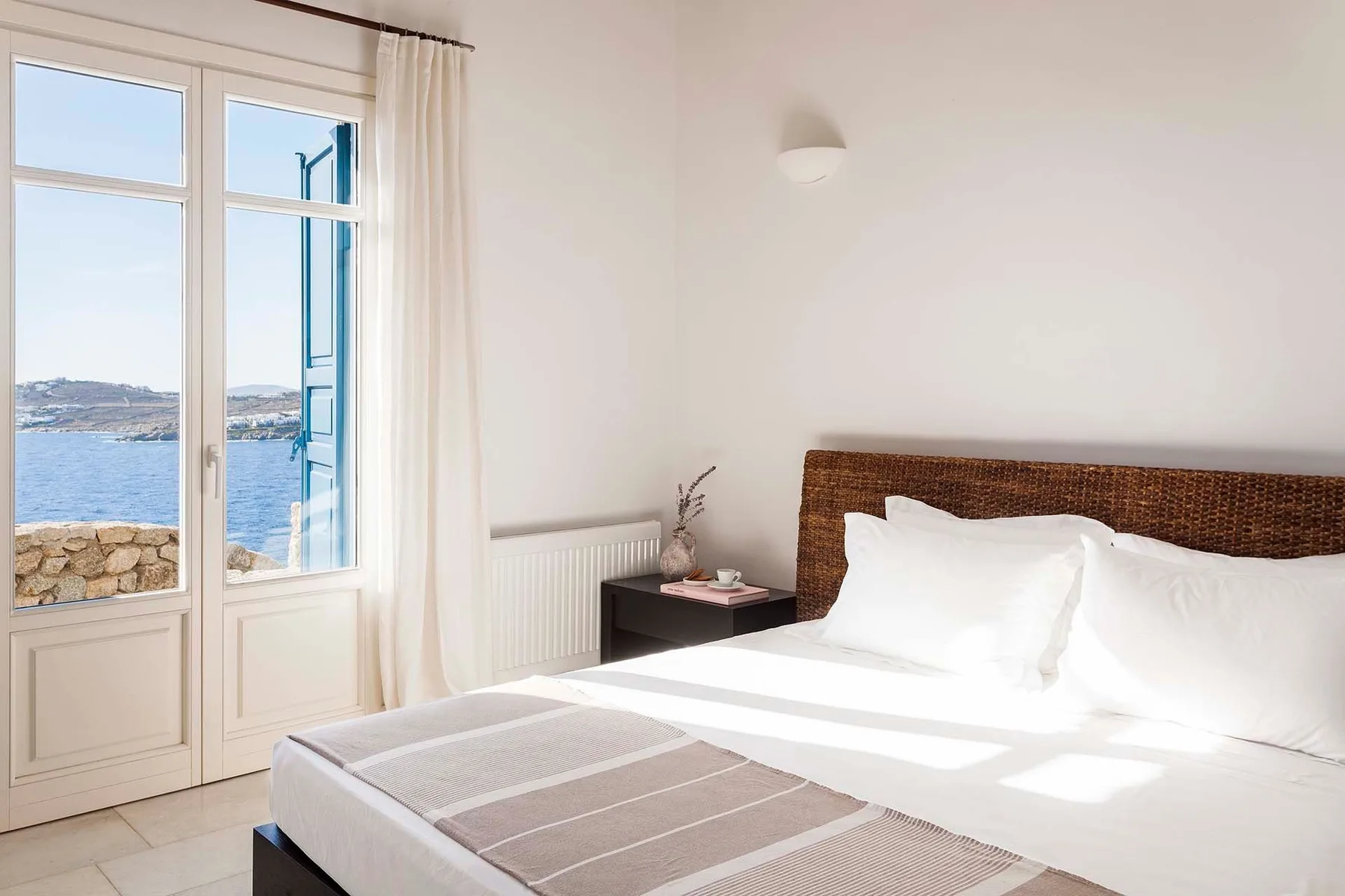A bright bedroom with a bed beside tall windows overlooking the water.