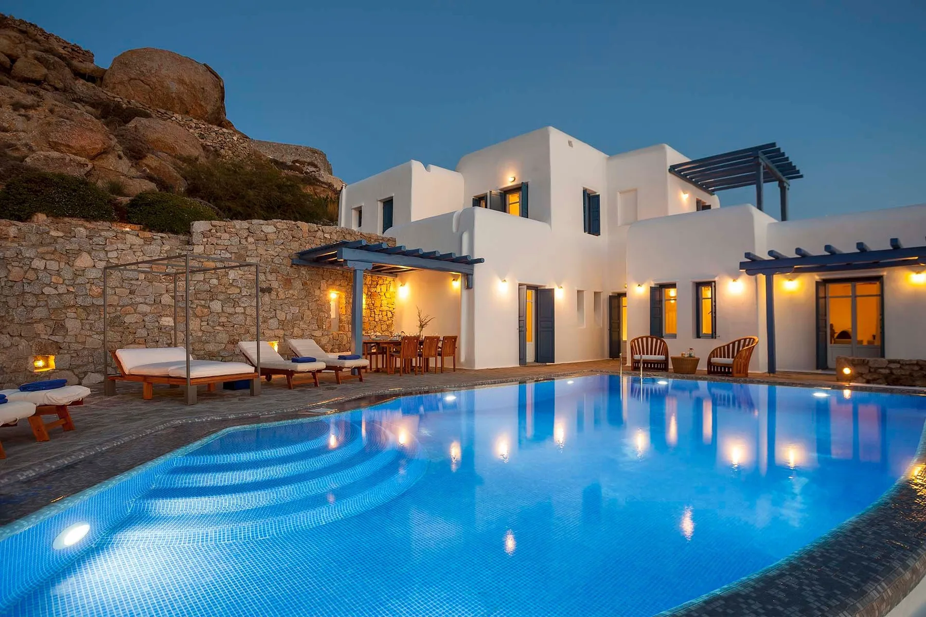 A white villa surrounds a lit swimming pool, with rocky terrain behind it.