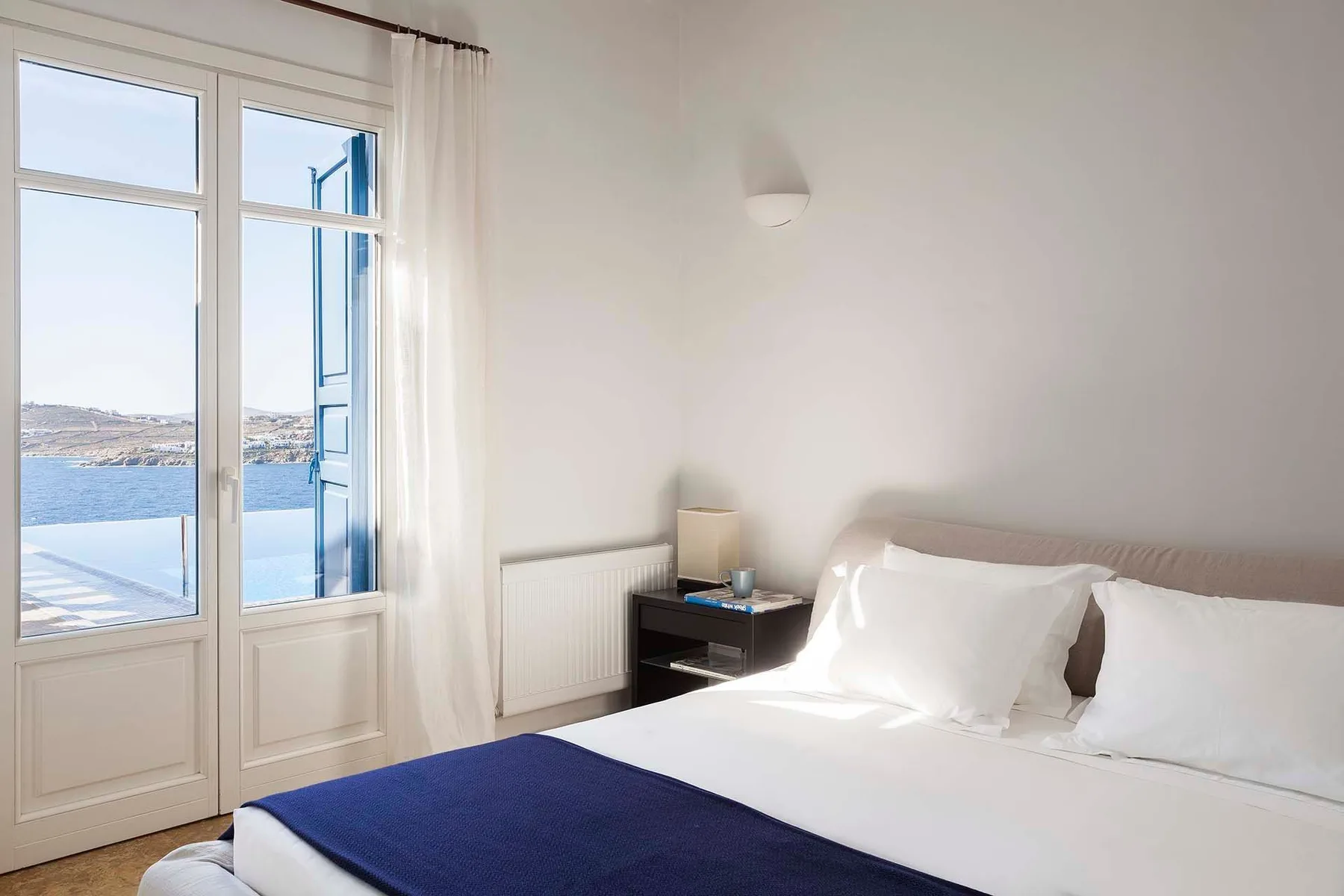A bright bedroom with a bed beside tall windows overlooking the water.