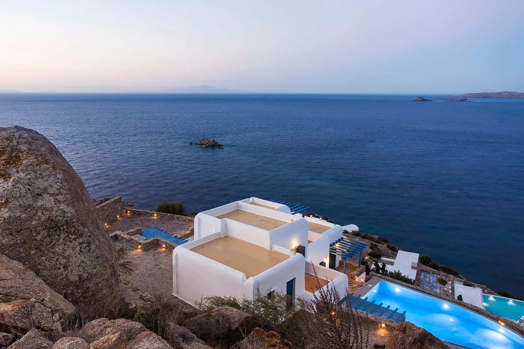 White buildings and illuminated pools sit beside the sea at dusk.