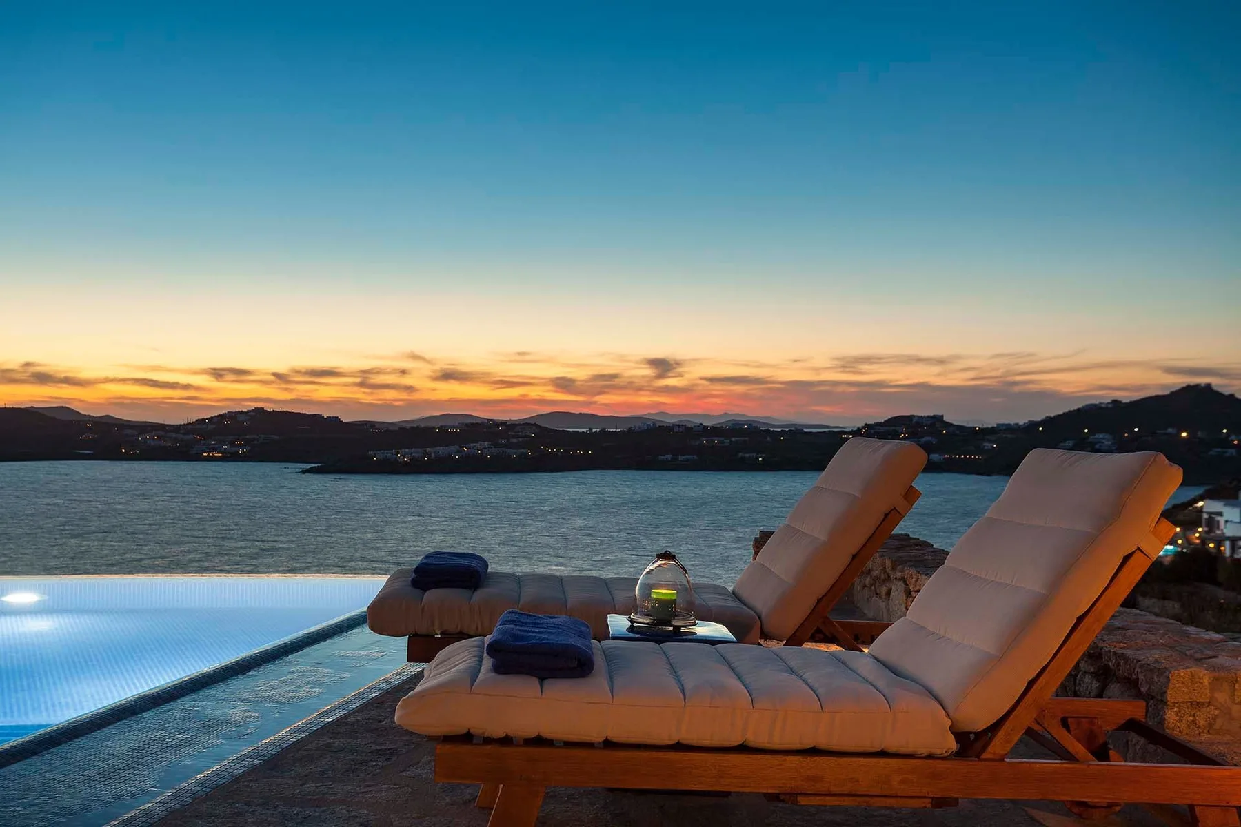 Poolside loungers overlook a calm bay beneath a colorful sunset sky.