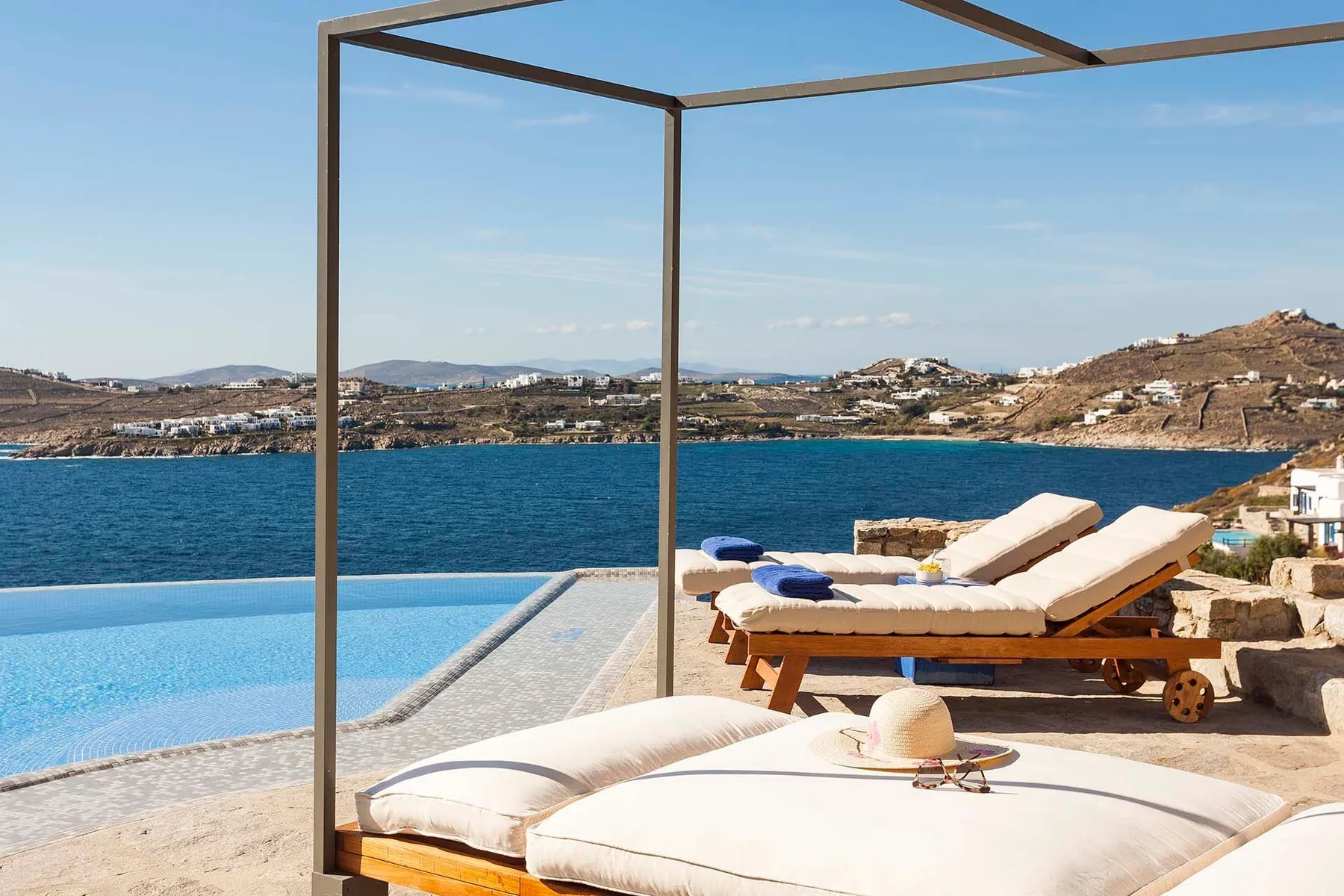 A poolside lounging area overlooks the sea, with cushioned seating beneath a pergola.