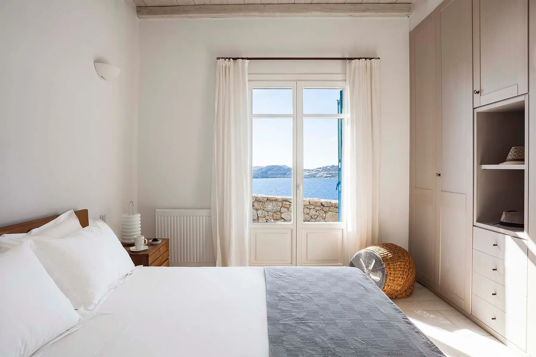 A bright bedroom with a bed beside a large window overlooking the water.