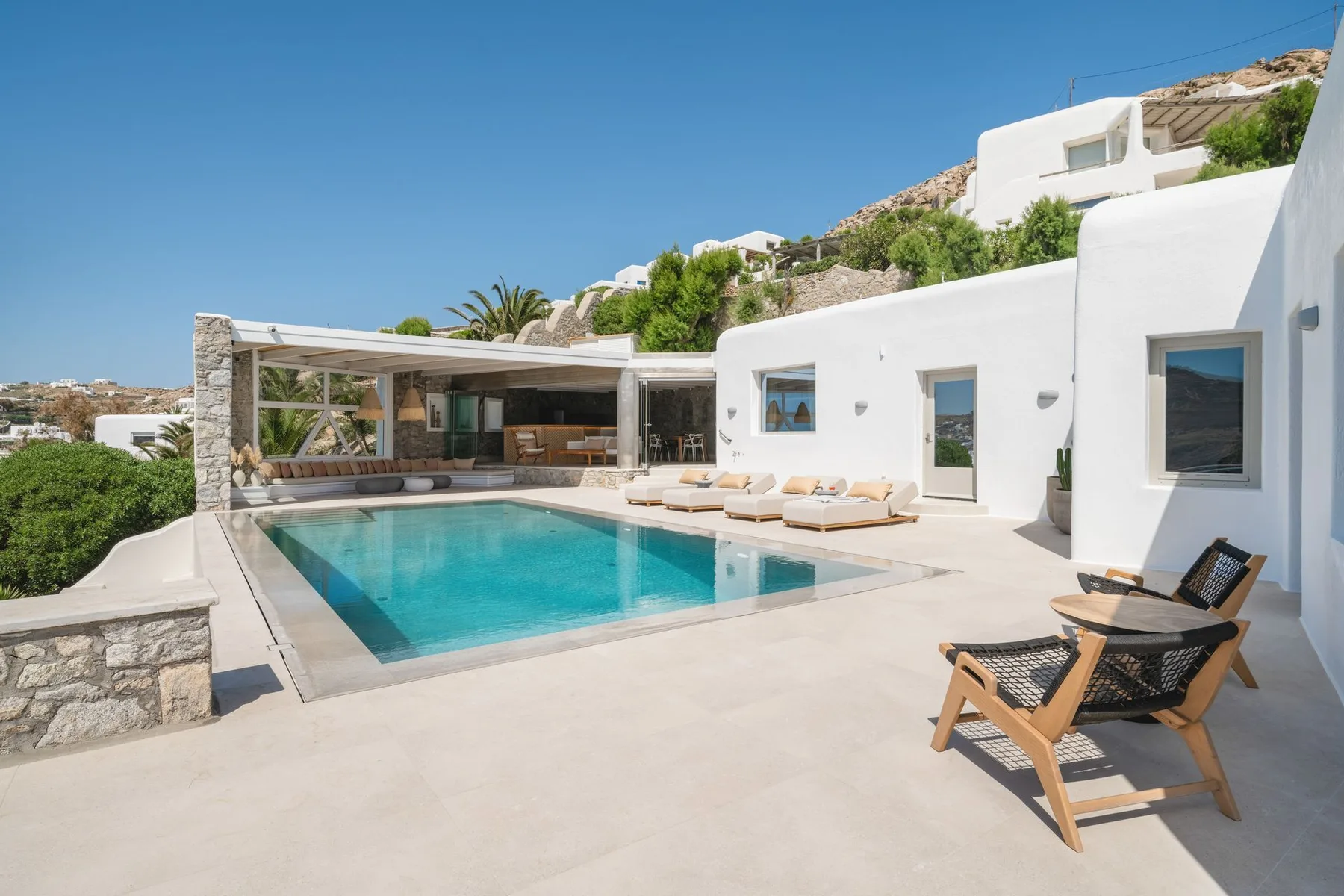 A sunlit courtyard features a swimming pool beside white buildings and outdoor seating.