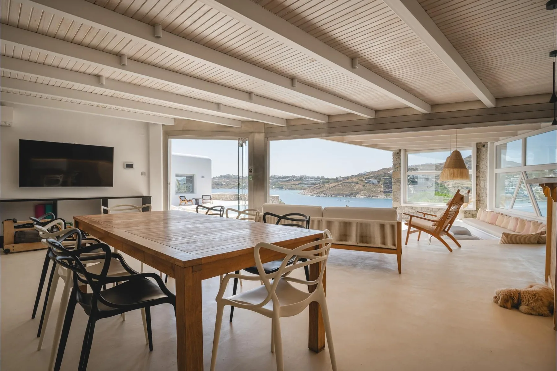A dining and living area with a wooden table and broad windows overlooking the water.