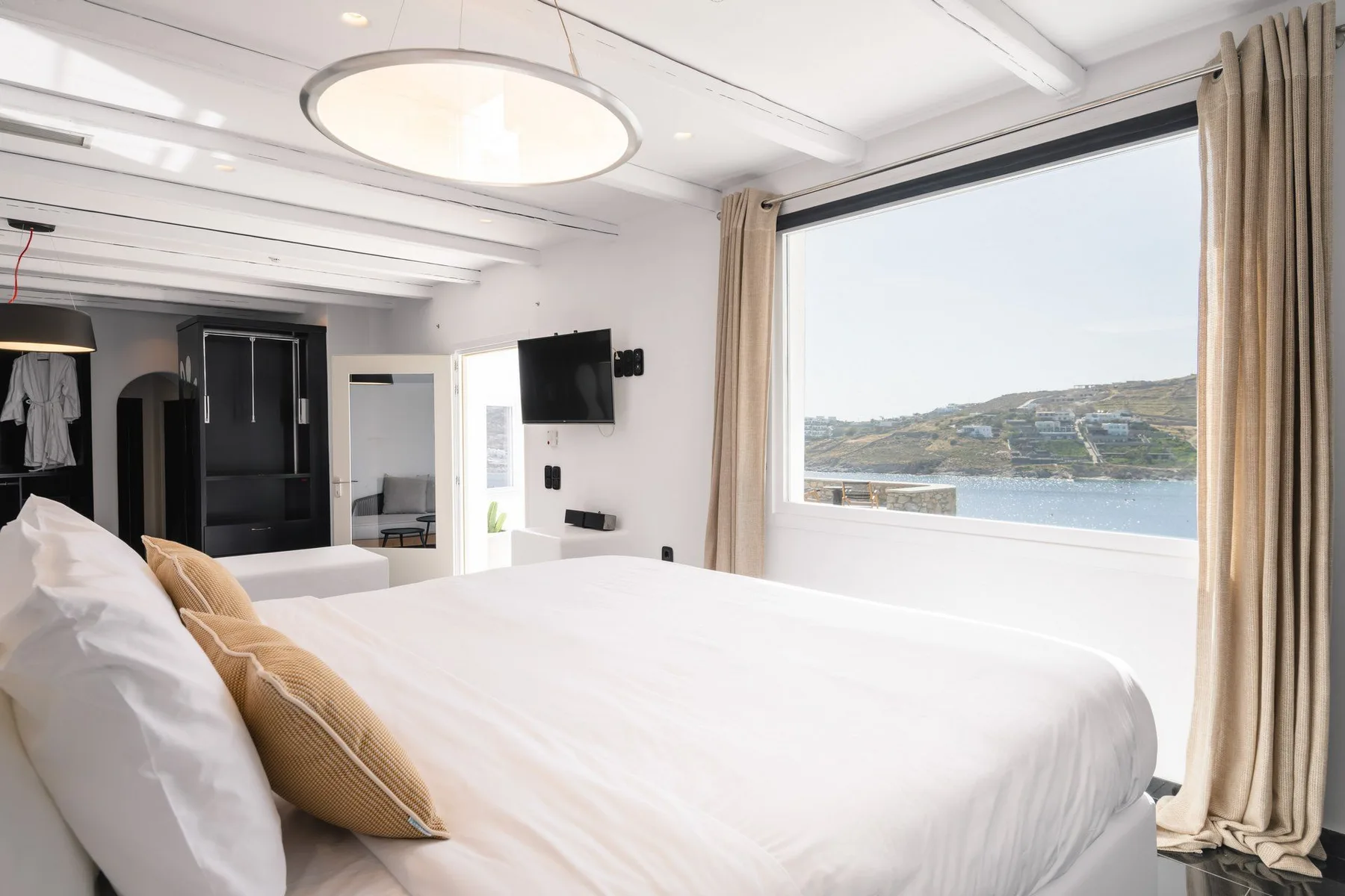 A bright bedroom with a large bed beside a wide window overlooking the water.