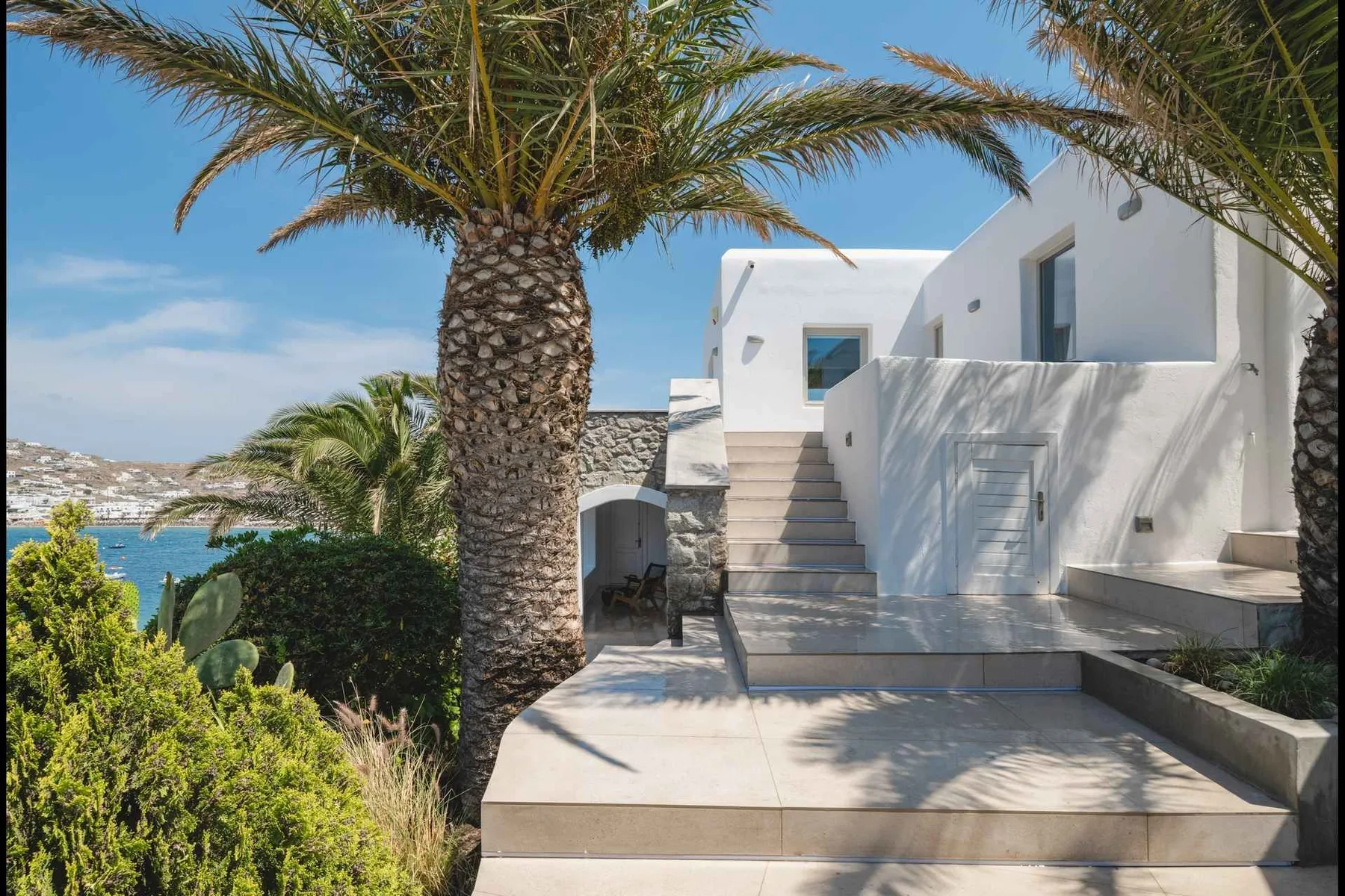 A white building with an outdoor staircase stands among palm trees overlooking the sea.