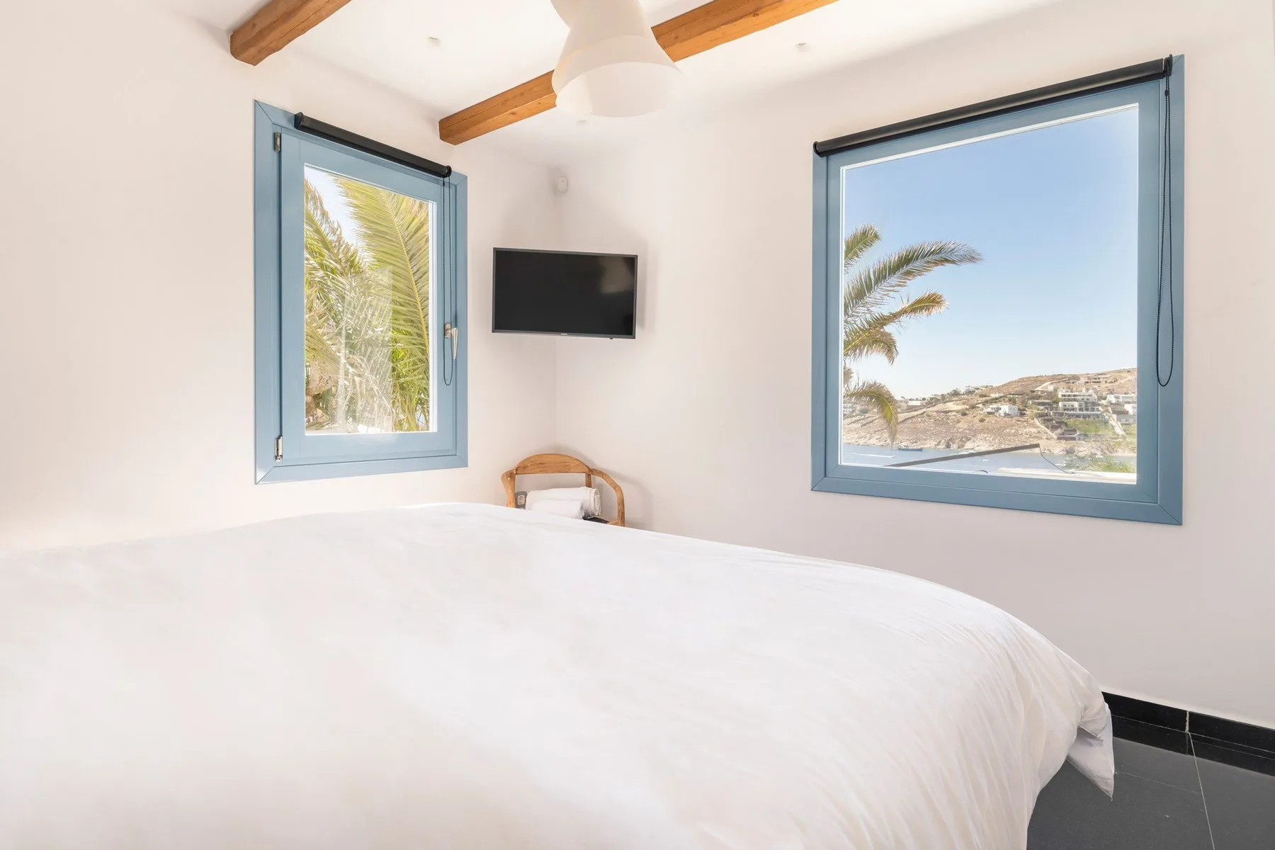 A bright bedroom with a white bed, blue-framed windows, and wooden ceiling beams.