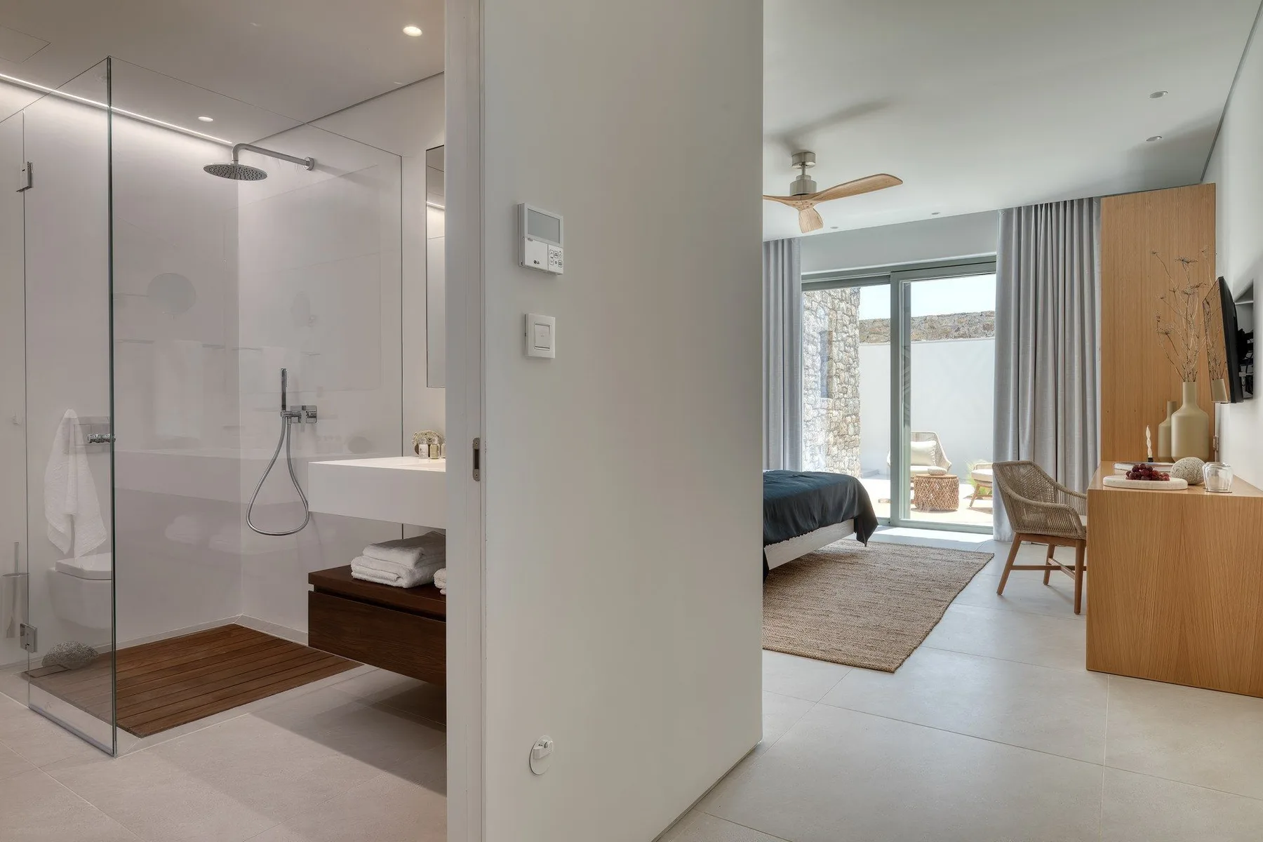 A bright bedroom opens onto a glass-walled shower area, with a bed and seating visible.