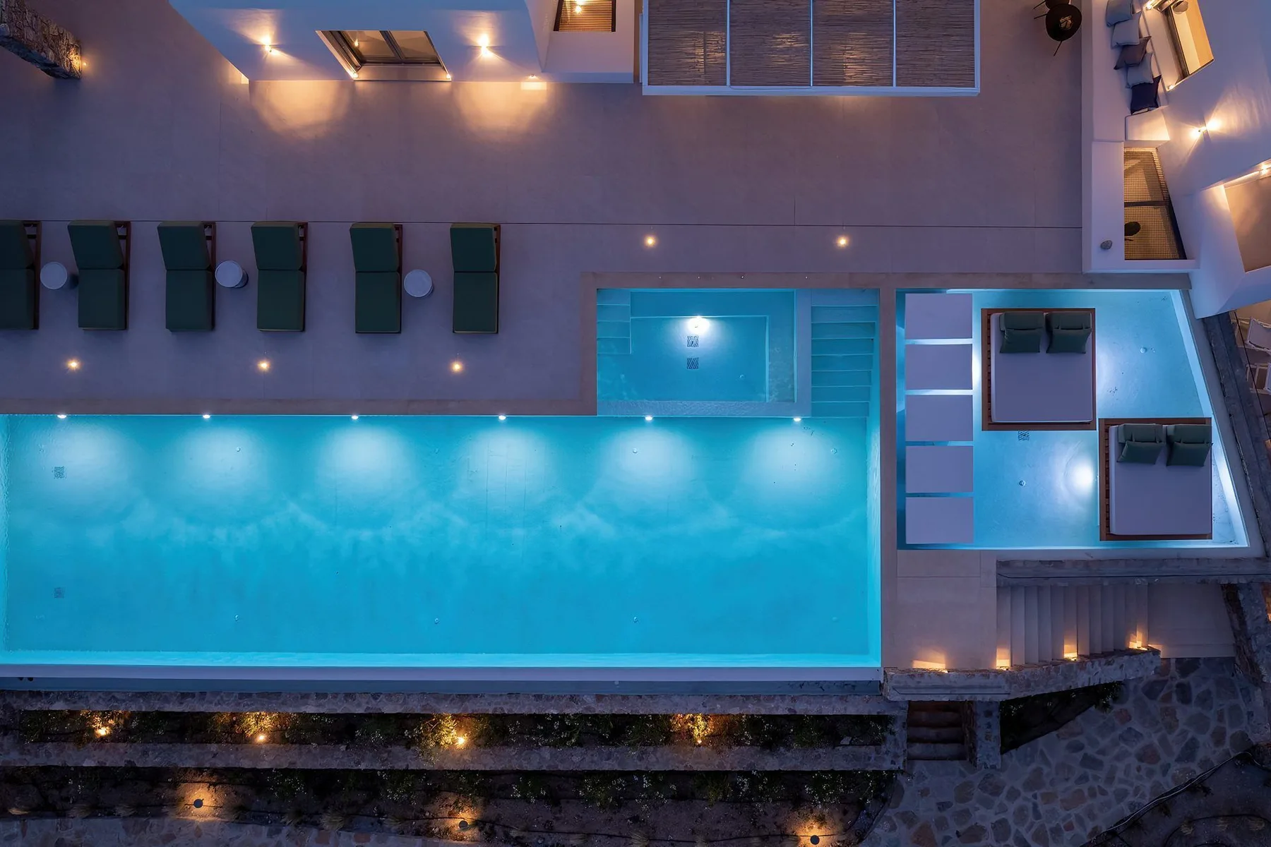 An illuminated swimming pool sits beside a paved patio with lounge seating.