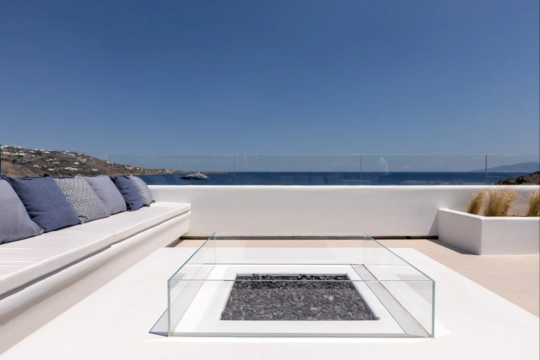 A sunlit seaside terrace with cushioned seating and a glass-covered feature.