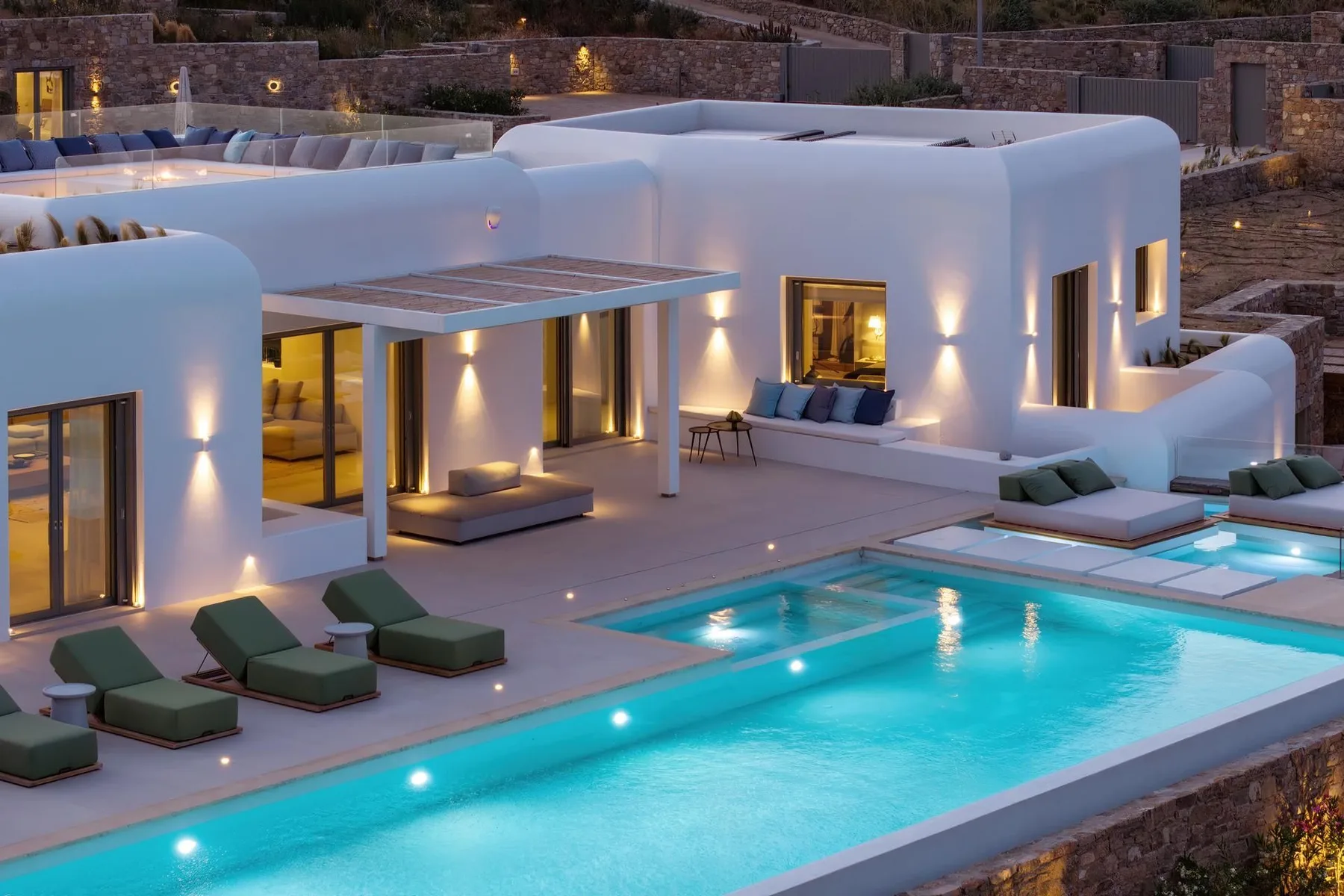 A modern white villa opens onto a lit swimming pool and outdoor seating area.