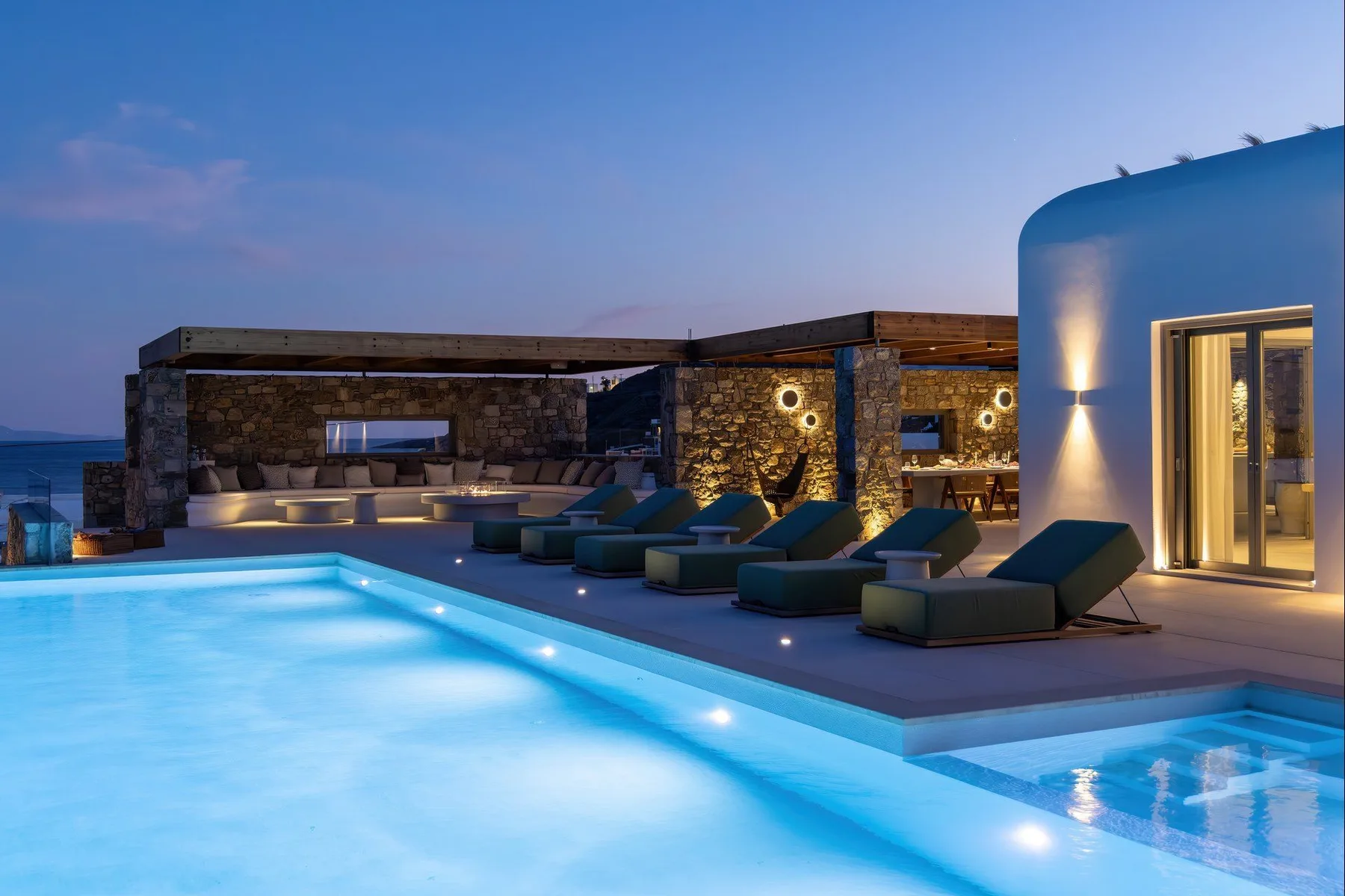 An illuminated outdoor pool sits beside lounge seating and a stone terrace at dusk.