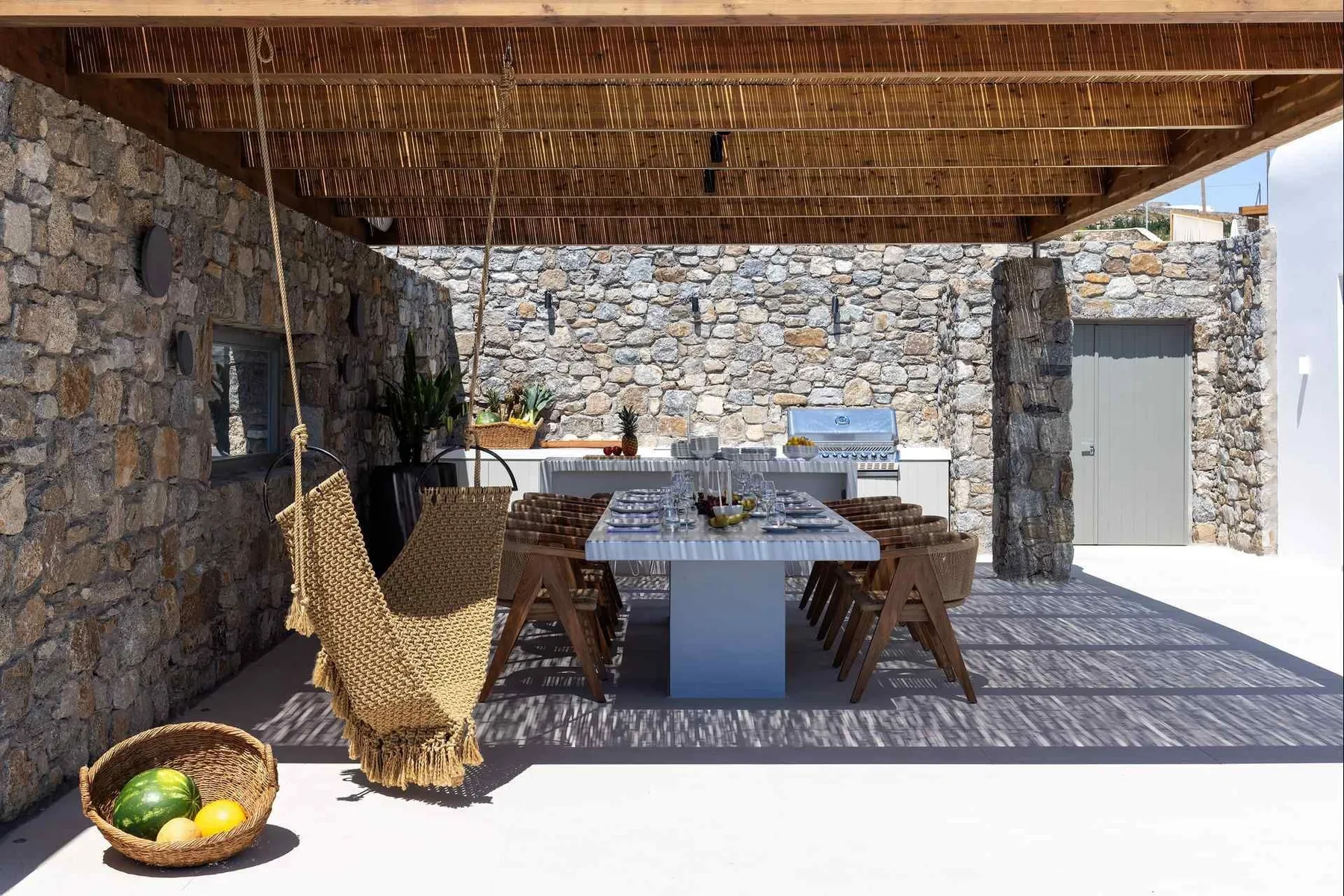 An outdoor dining area sits beneath a wooden pergola, with a long table and stone walls.