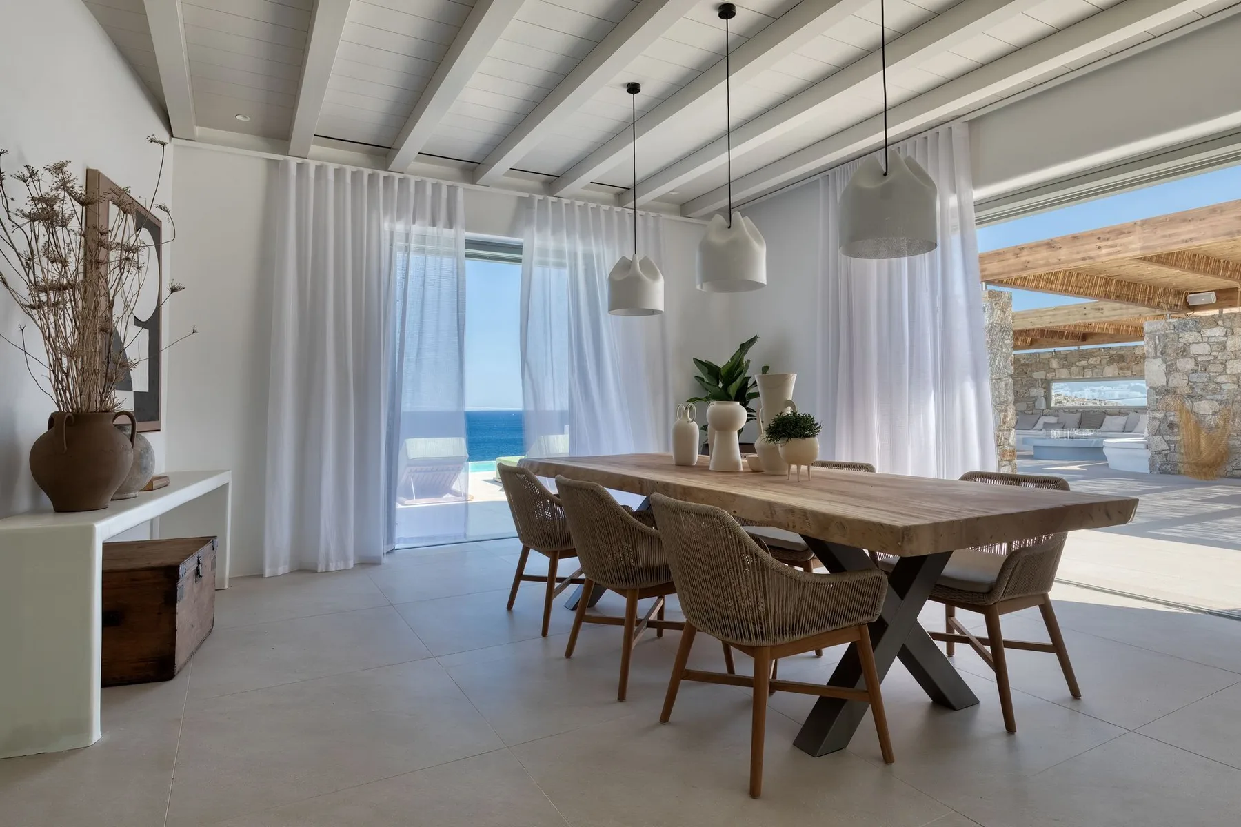A bright dining area with a long table beside large windows overlooking the sea.