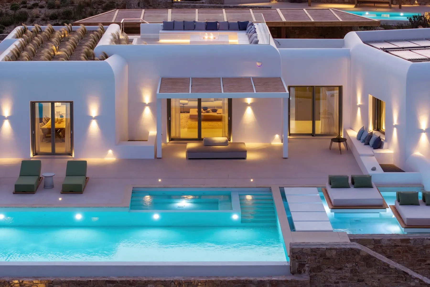 A modern white house opens onto a lit swimming pool and patio at dusk.