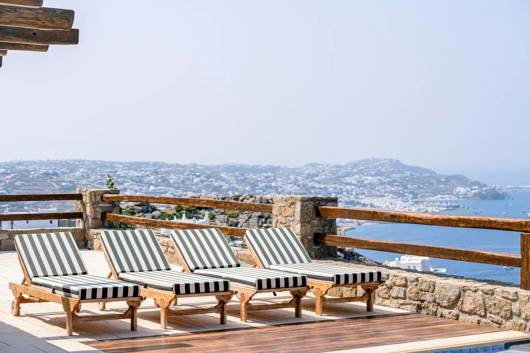 A sunlit terrace with striped loungers overlooks the sea and distant hills.