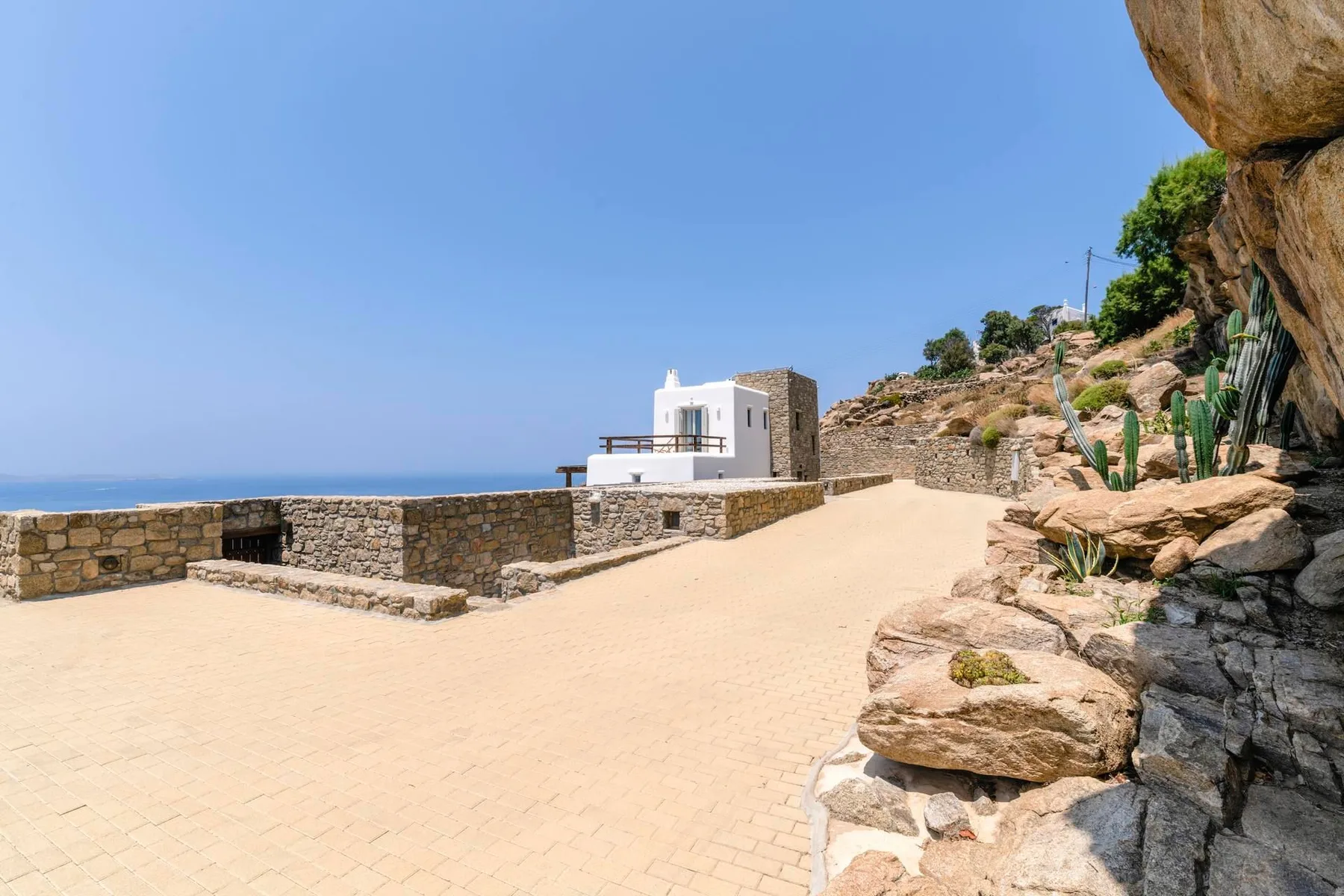 A sunlit stone terrace beside a white building overlooks the sea.