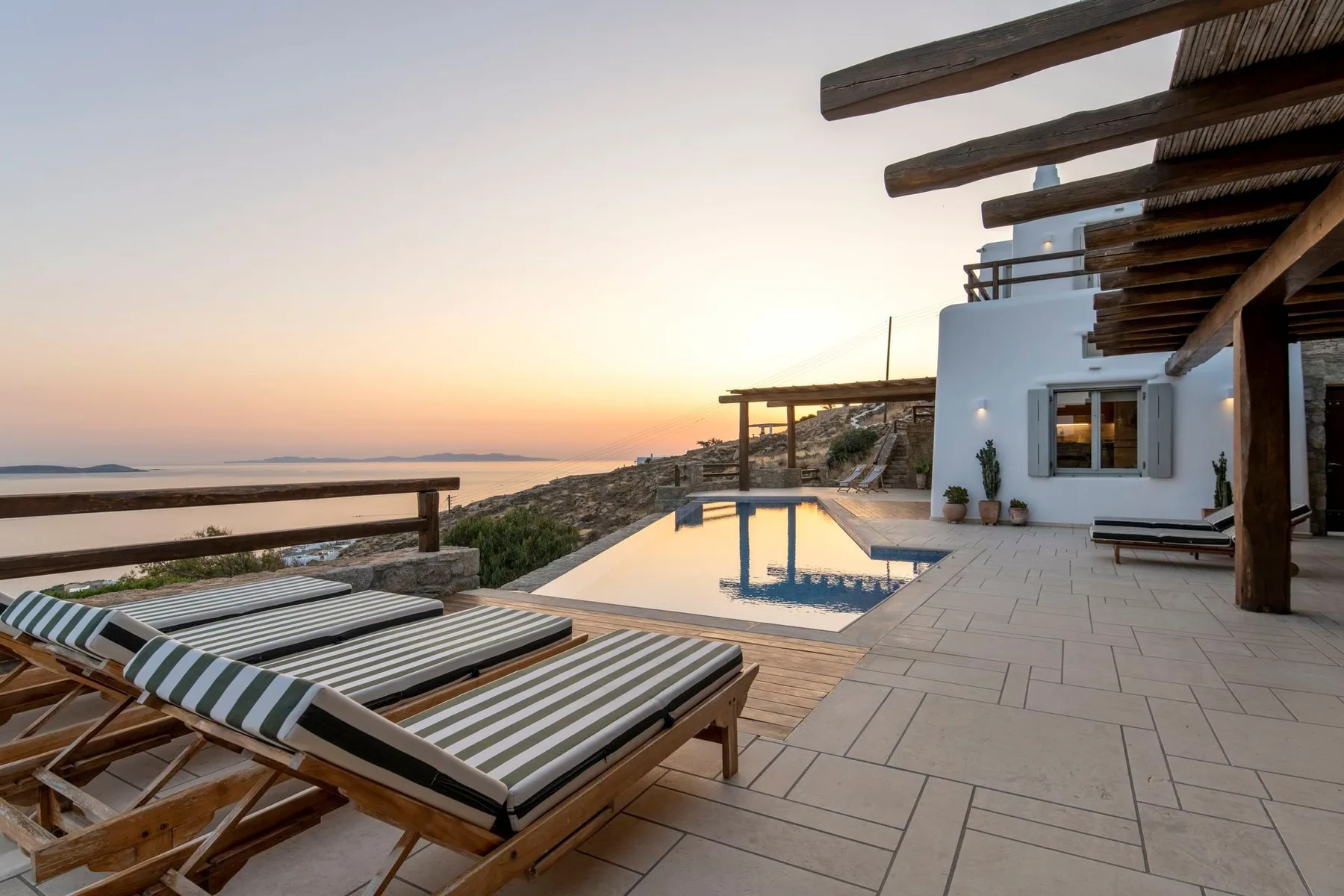 A seaside terrace with striped loungers beside a swimming pool at sunset.