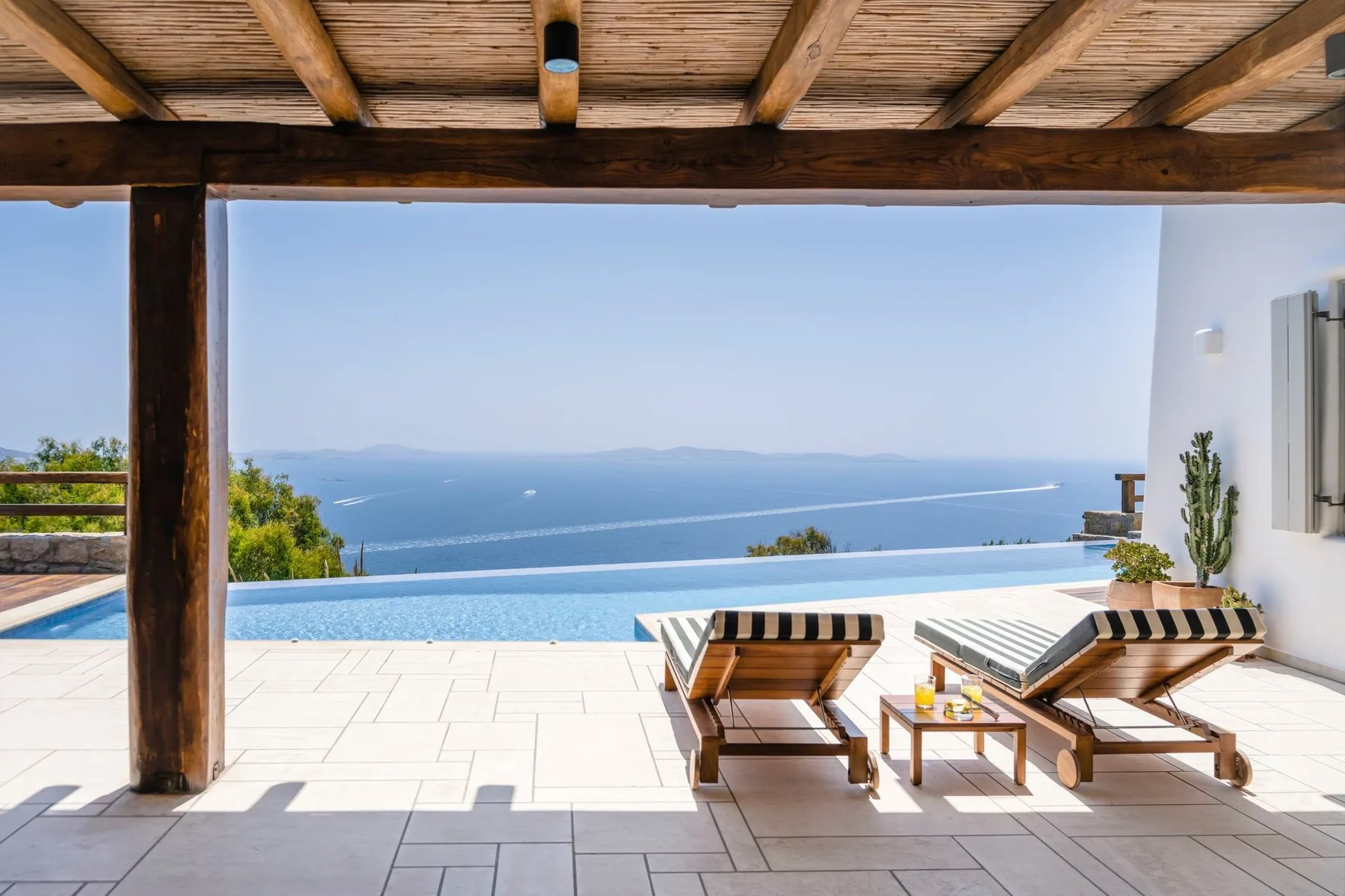 A shaded patio overlooks a swimming pool and the sea, with loungers nearby.