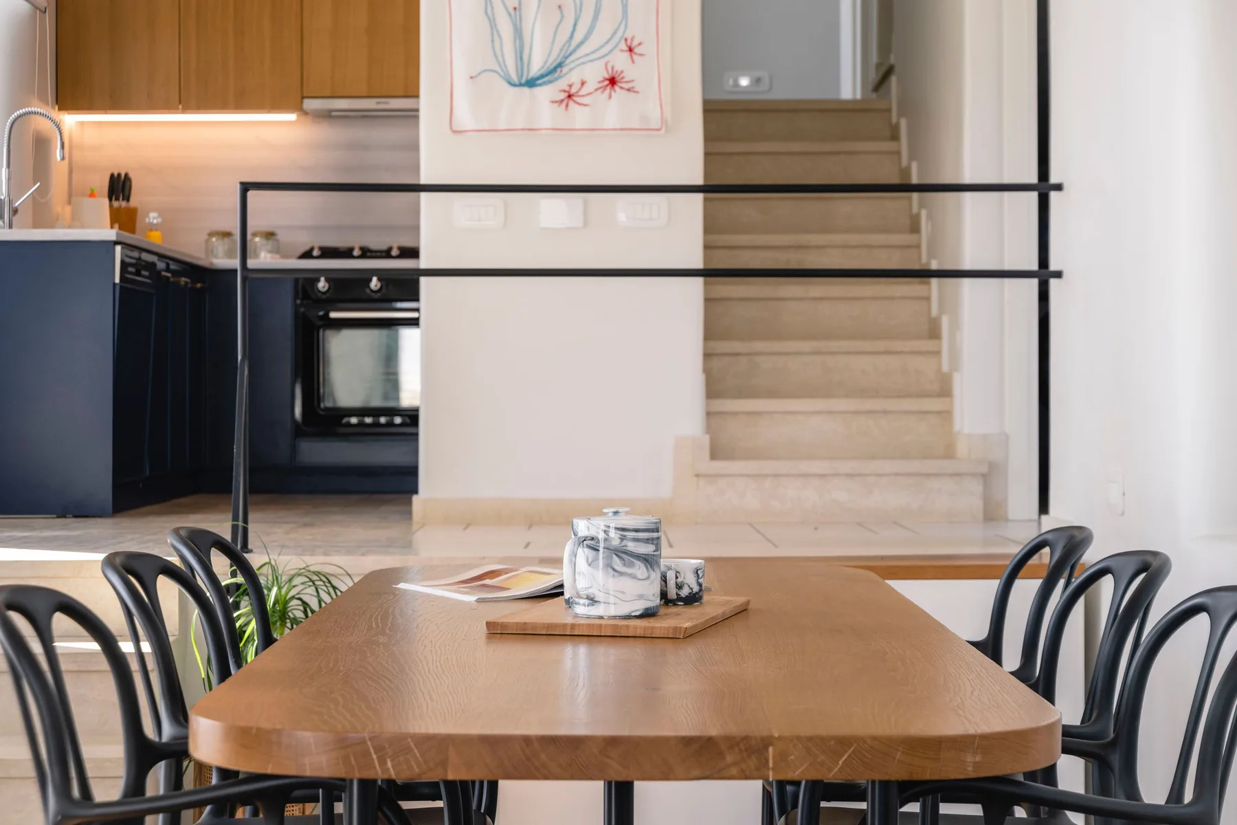 A dining table and chairs sit in front of a kitchen and staircase.