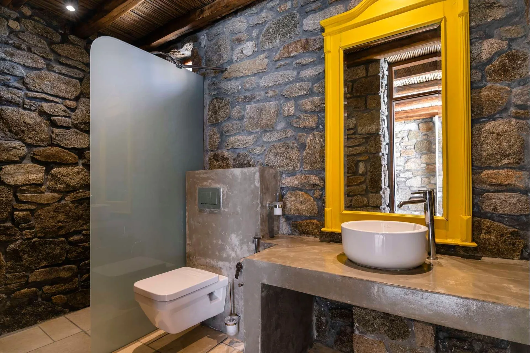 A stone-walled bathroom with a sink and toilet beneath a wooden ceiling.