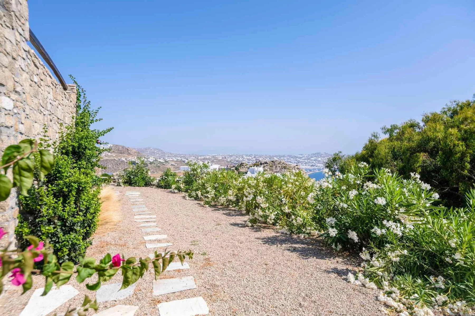 A gravel garden path runs between flowering shrubs, with a distant landscape view.