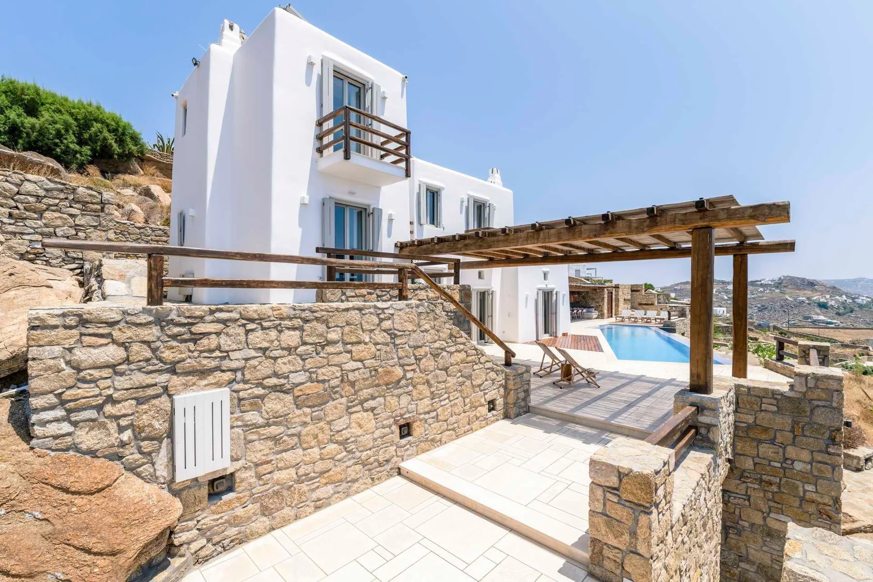 A white building and outdoor pool sit beside a stone terrace and wooden pergola.