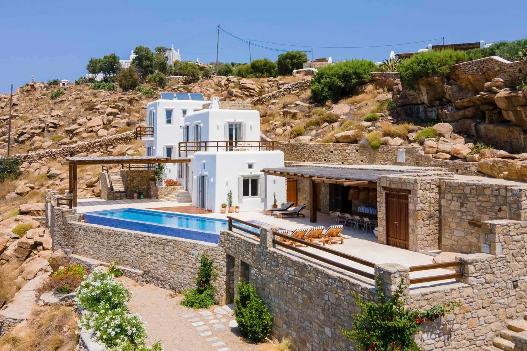 White buildings sit on a rocky hillside beside a swimming pool and stone terrace.