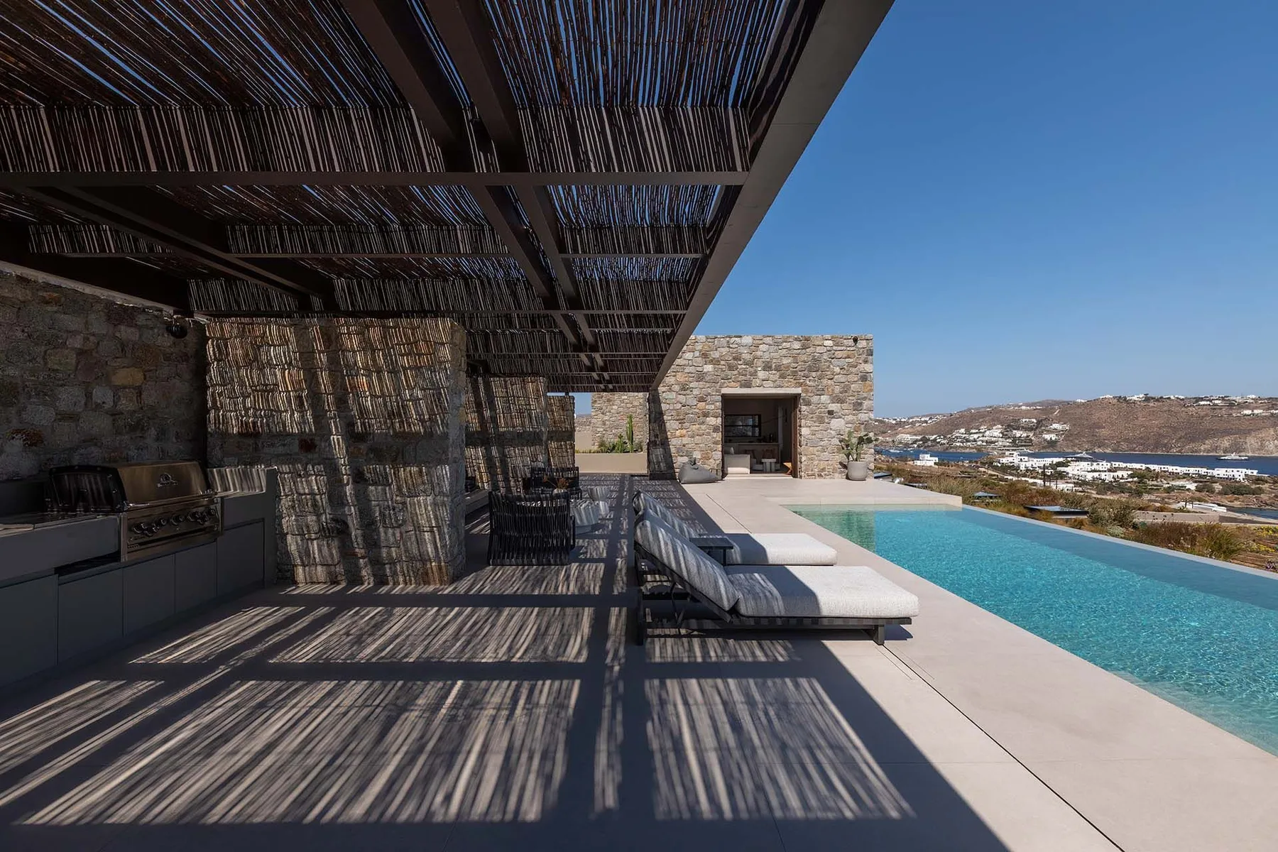A shaded outdoor kitchen and seating area overlooks a swimming pool and distant hills.