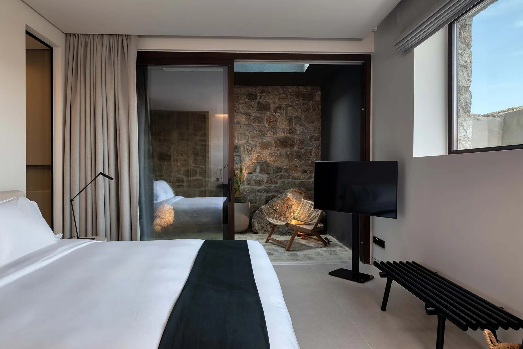 A bedroom with a bed, a television, and a seating area beside a stone wall.